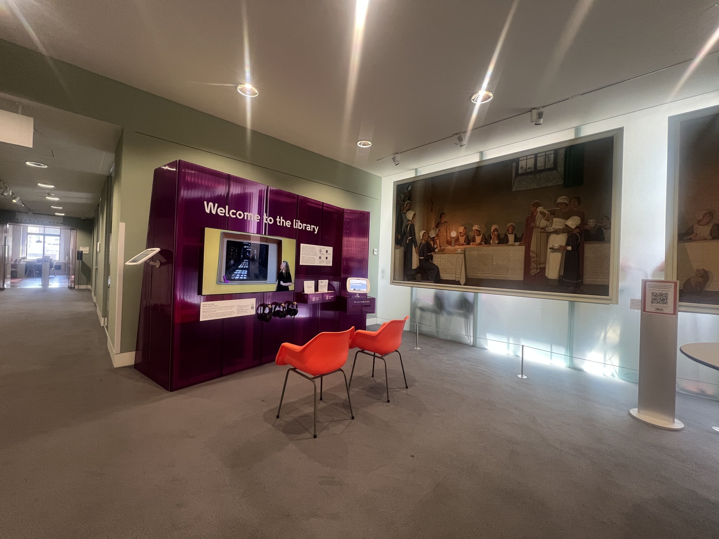 Interior of a museum or library with a mural of historical figures, a purple information kiosk, and orange chairs.