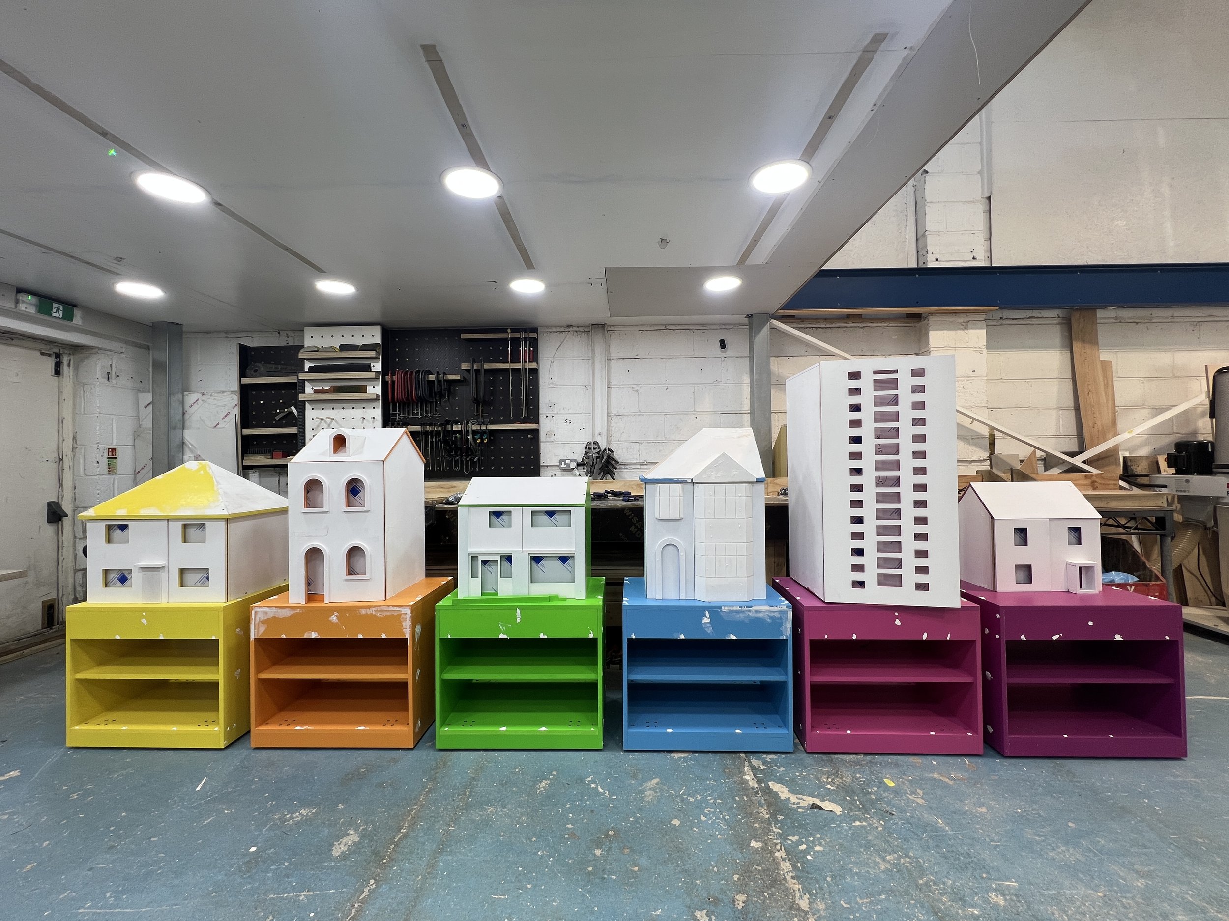 Colorful display of small painted wooden dollhouses arranged in a row in an indoor workshop space.