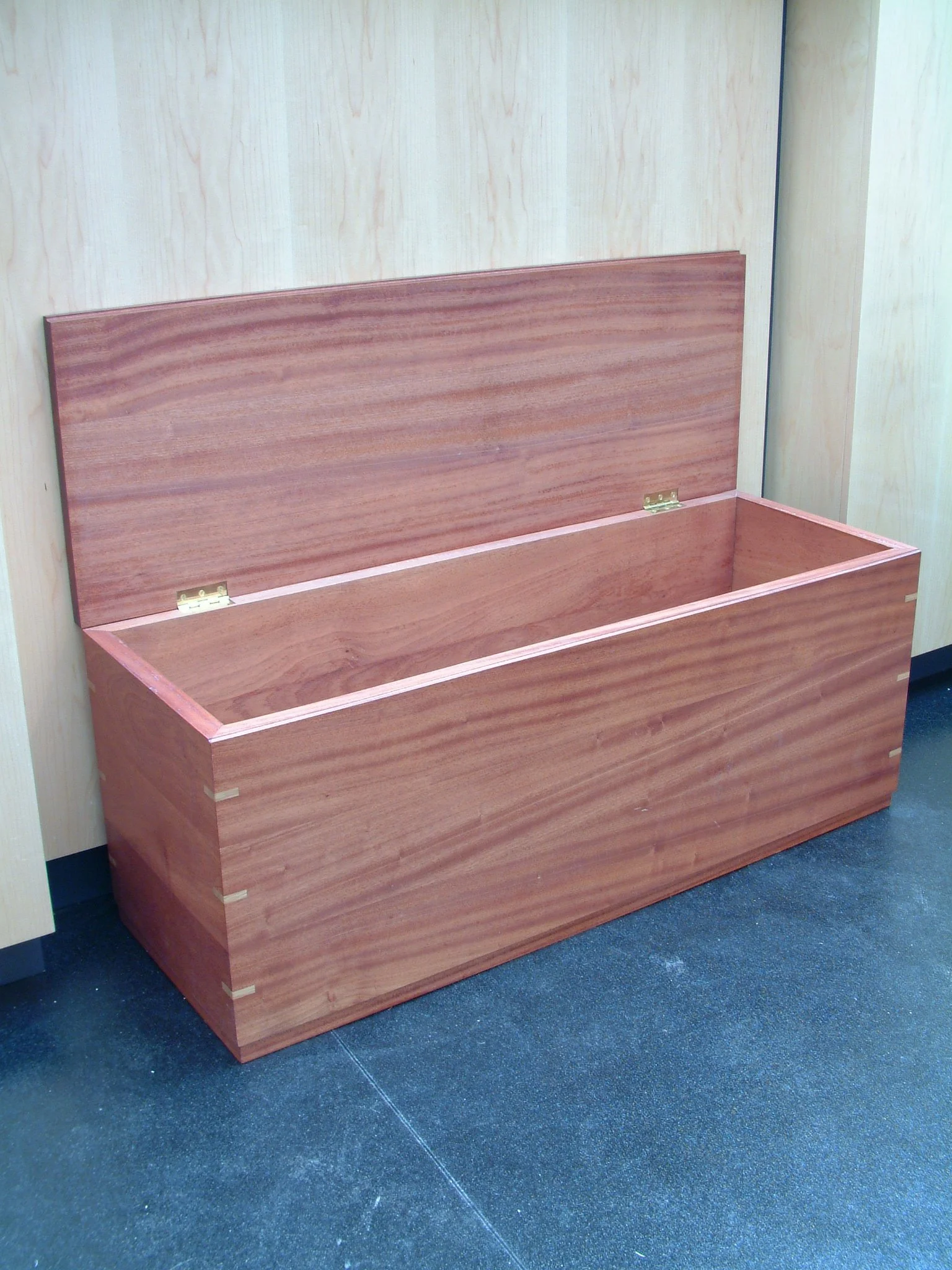 Wooden storage chest with open lid, showing empty interior, placed on a dark blue carpeted floor against light-colored wood-paneled wall.