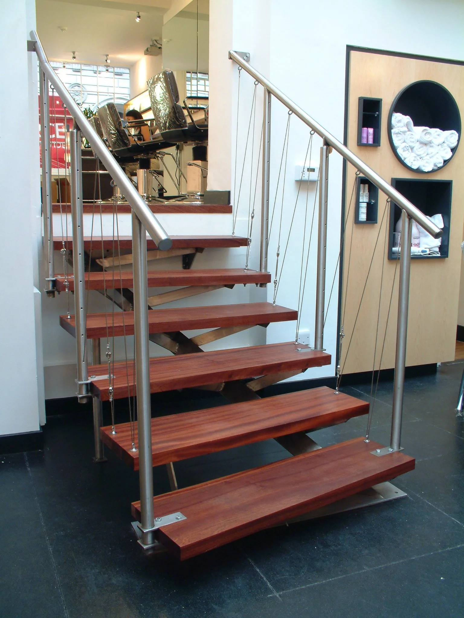 A modern staircase with wooden steps, steel handrails, and cable supports, located inside a building with black flooring and a wall-mounted display containing rolled towels and small shelves.