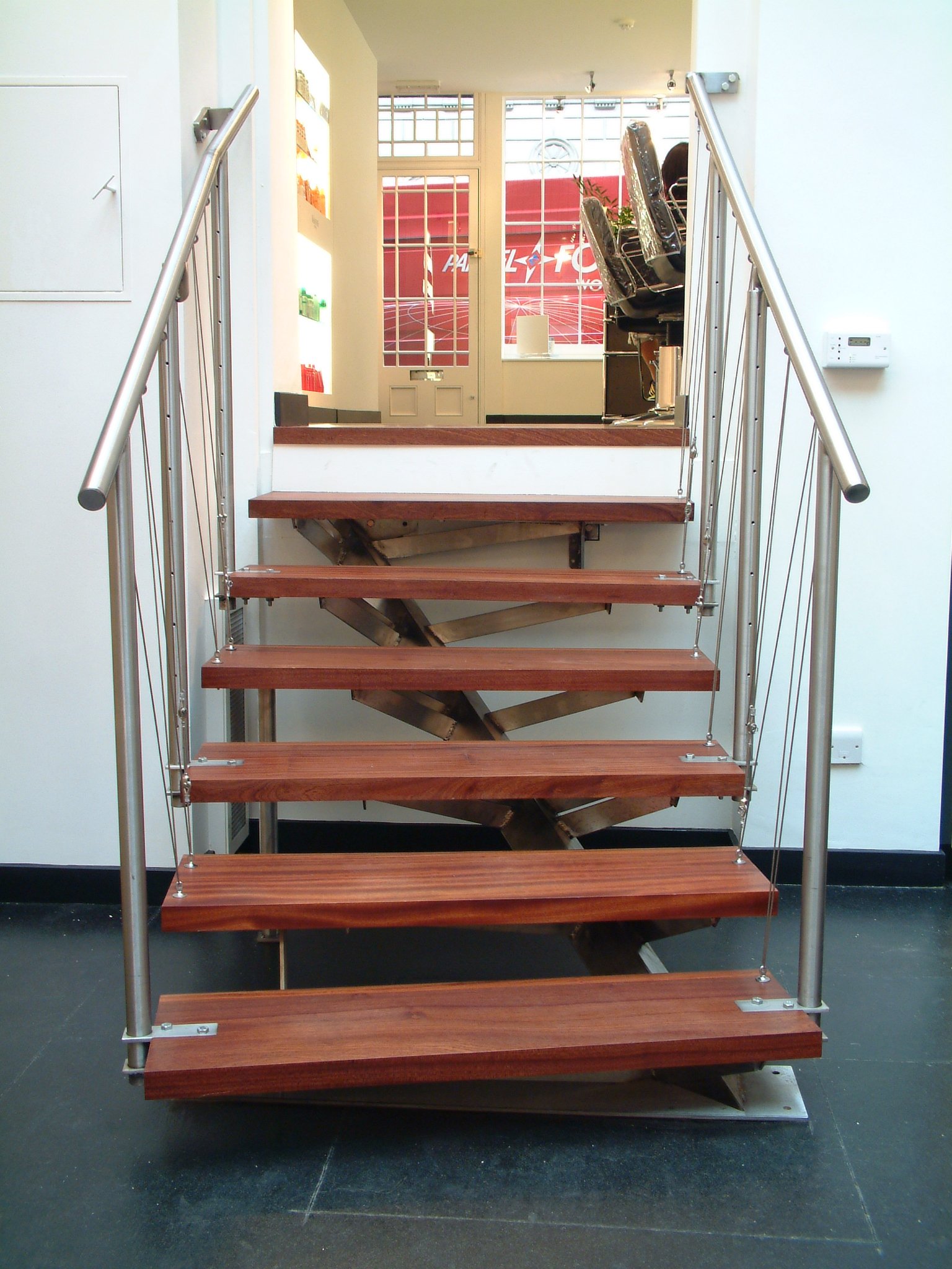 A modern staircase with wooden steps and metal railings, leading up to an entryway with glass doors and a red exterior visible outside.