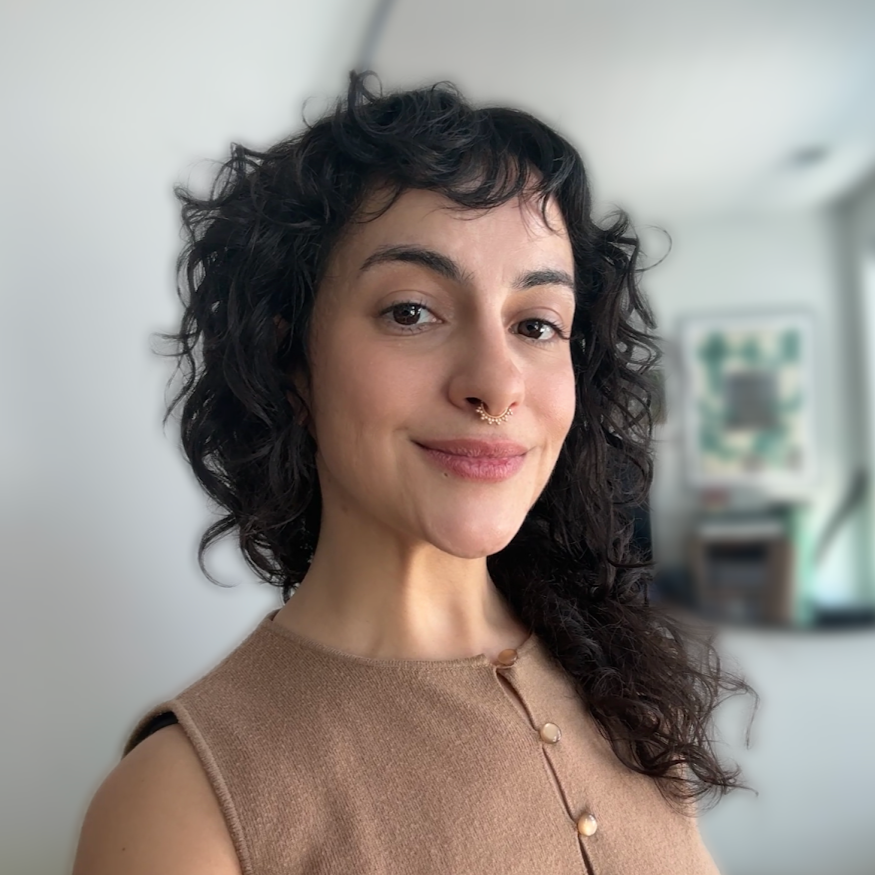 A woman with curly dark hair, a septum piercing, wearing a sleeveless beige top, smiling in a bright room.
