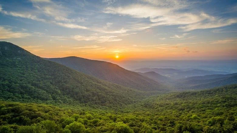 Sunset over a mountain range with green forested hills and a sky with clouds.