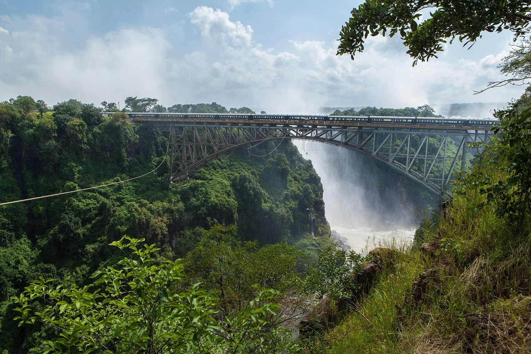 A large bridge spans a lush, green, forested canyon with a waterfall below. A train is crossing the bridge, and mist rises from the waterfall.