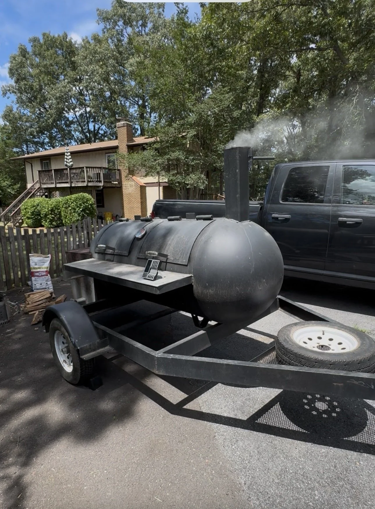 A black barbecue smoker on a trailer with smoke coming out, parked in a driveway next to a black truck, with a house and trees in the background.