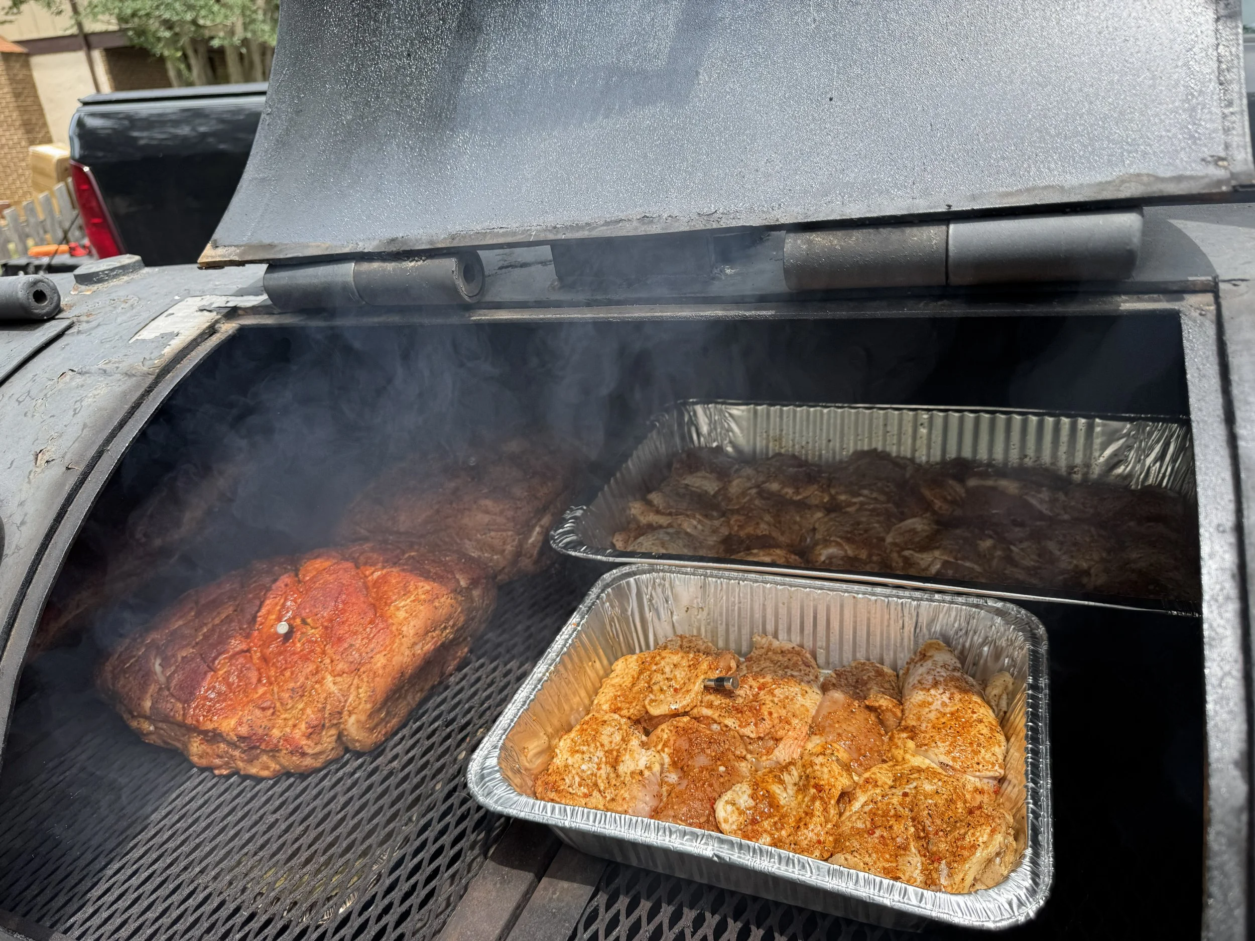 Outdoor cooking setup with a grill and several trays of seasoned raw and cooked meat, including chicken and beef, with smoke rising.