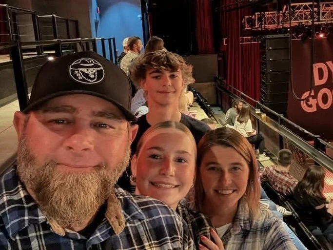 Four people taking a selfie at an indoor event or concert, with a stage and audience seated in the background.