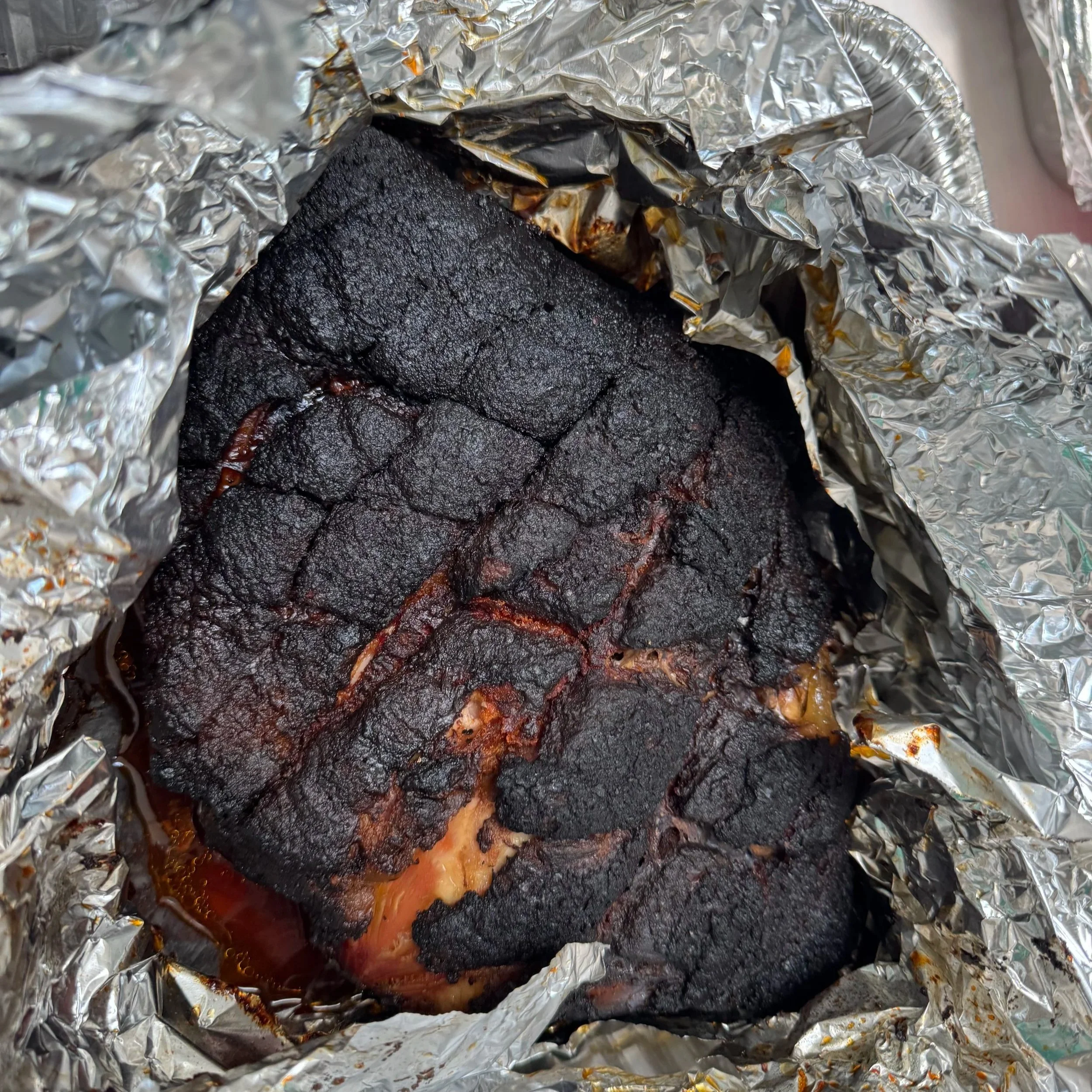 Charred, burnt meat wrapped in aluminum foil, with visible blackened surface and some juices seeping out.