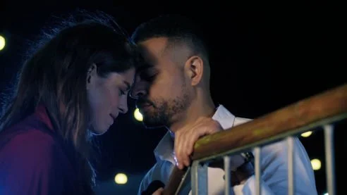 A man and woman close together with foreheads touching on a balcony at night.