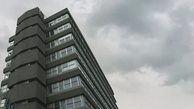 A tall concrete apartment building with multiple stories, seen from below against a cloudy sky.