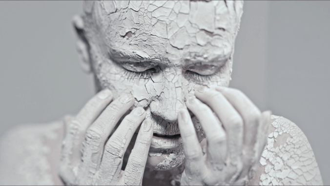 A person with cracked white clay or paint on their face and hands, holding their face with their eyes closed.