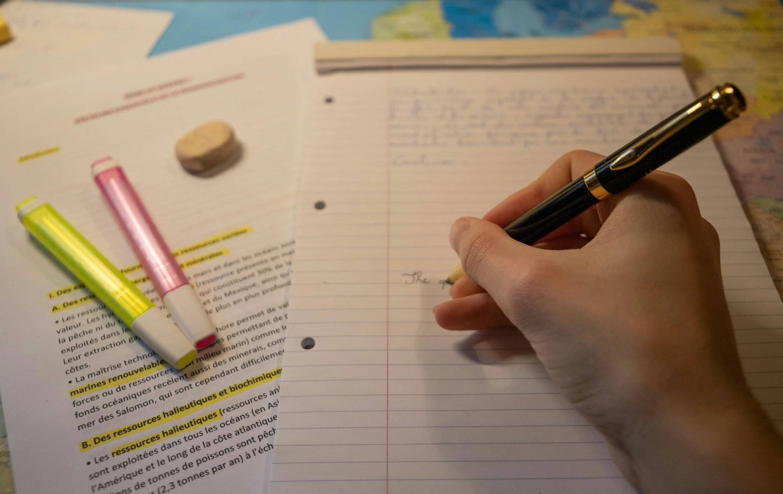 A person writing in a lined notebook with a black pen, placed on top of a map. There are two highlighter markers, one yellow and one pink, and some printed pages with highlighted text underneath the notebook. A small eraser is also on the pages.