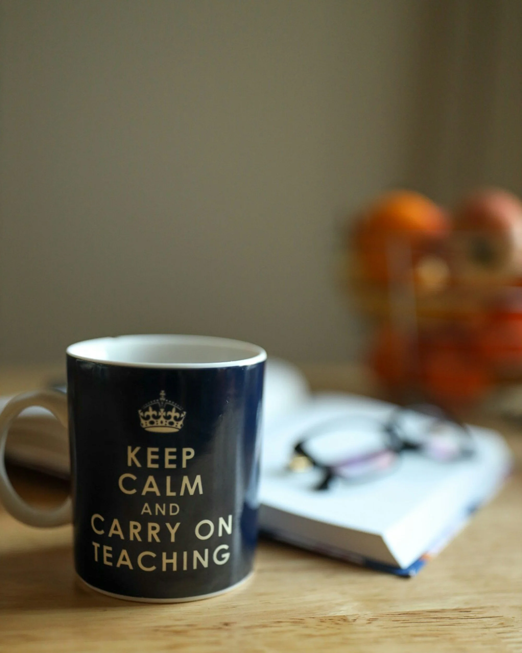 A blue coffee mug with a design that reads, "KEEP CALM AND CARRY ON, TEACHING," placed on a wooden surface with blurred objects and papers in the background.