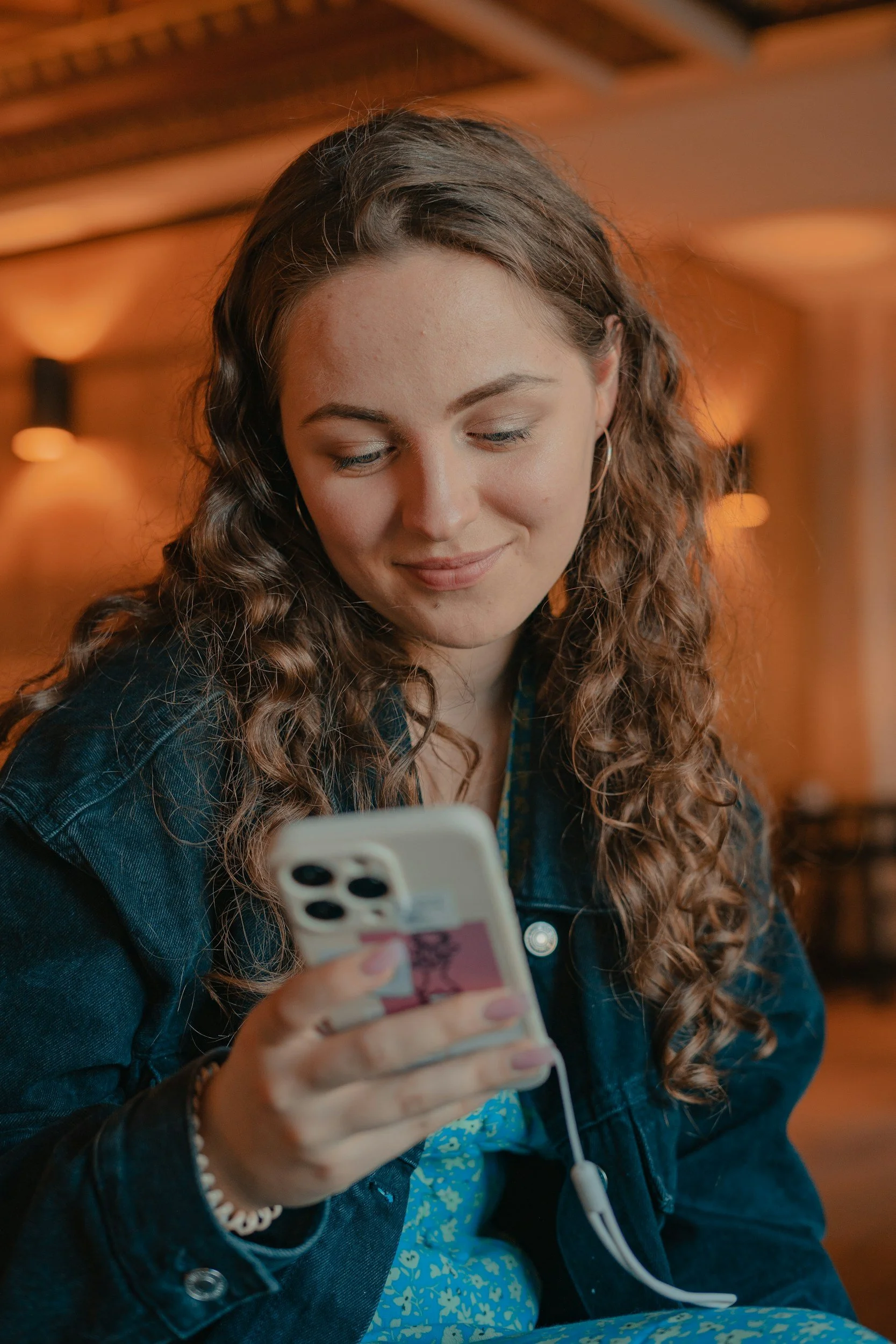 A young woman with curly brown hair looking at her phone with a smile in a warmly lit indoor setting.