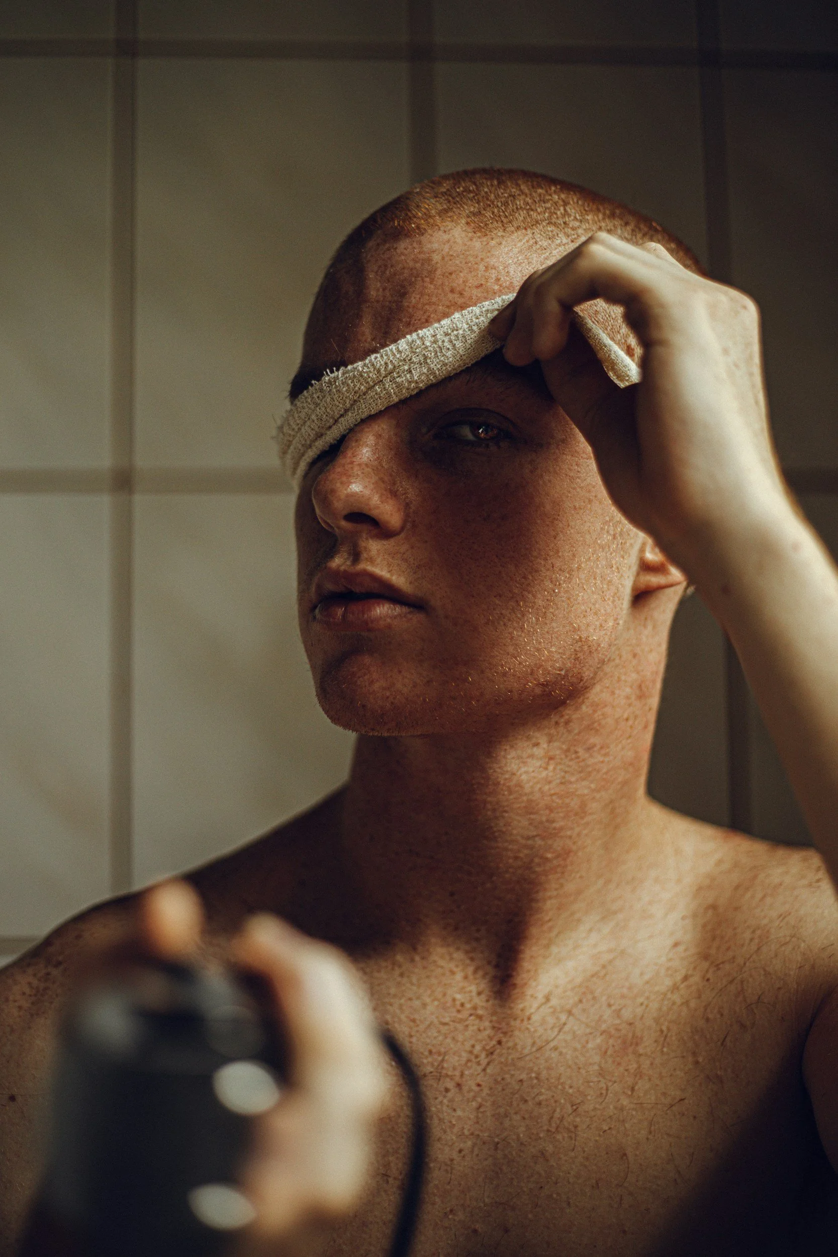 A person with short hair and freckles is holding a mirror, partially covering their face with a cloth, and looking into the mirror while aiming a gun towards the camera.