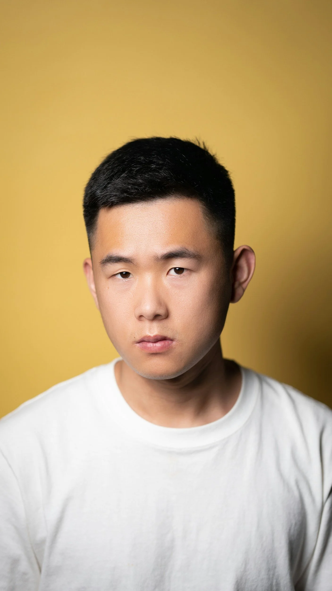 Portrait of a young Asian man with short black hair, wearing a white T-shirt, against a yellow background.