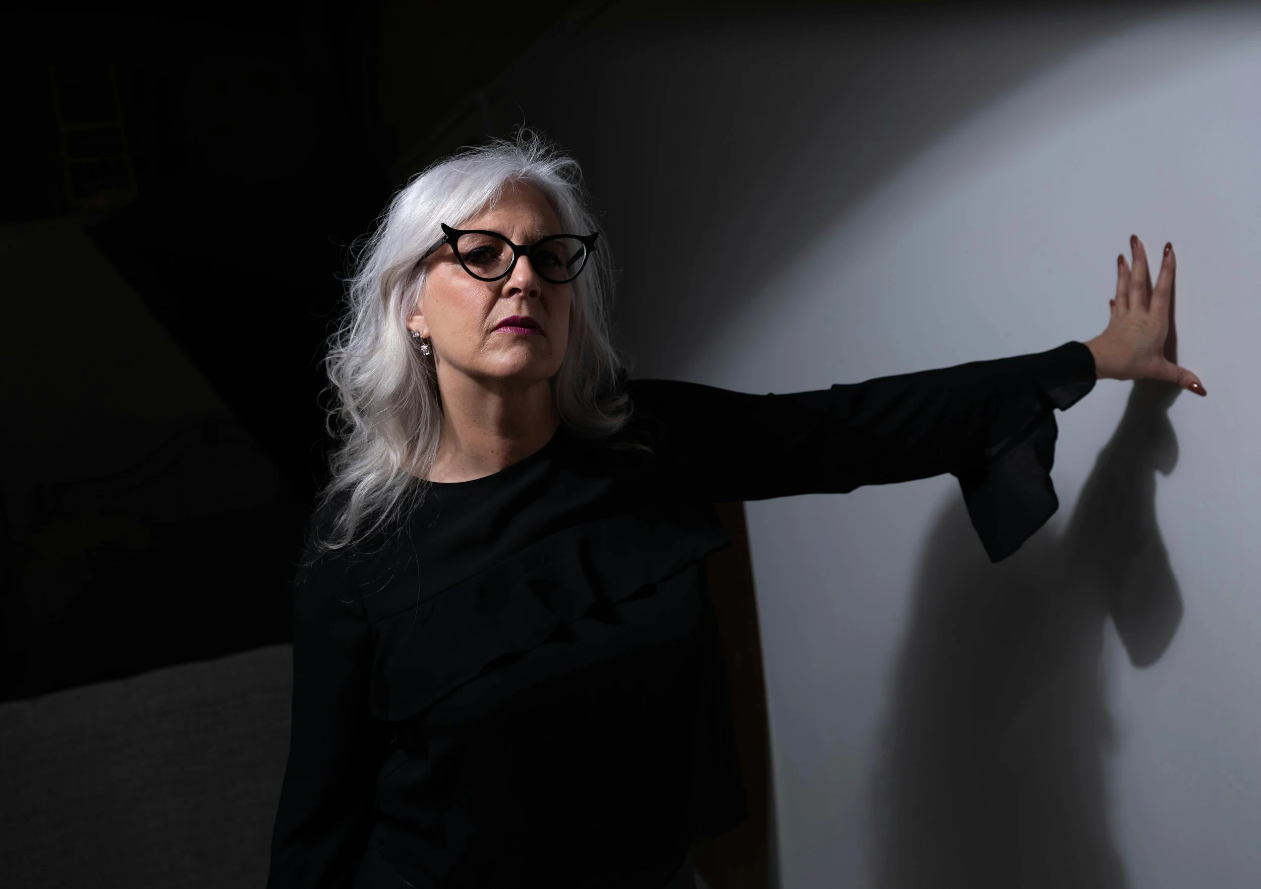 A woman with white hair wearing glasses and dark clothing, with her arm extended touching a plain wall.