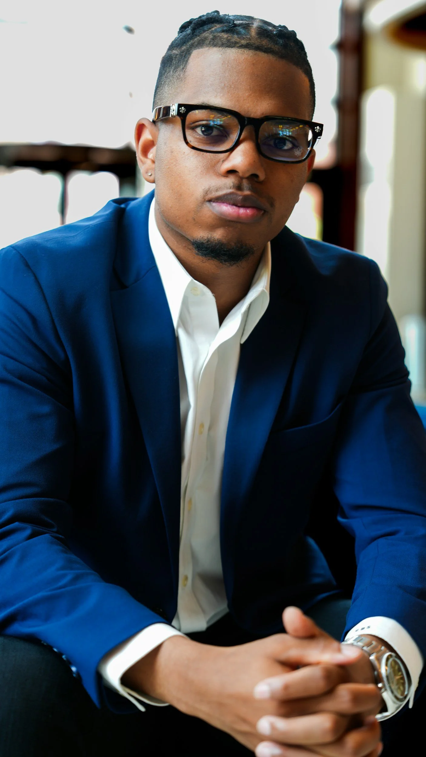 A young man with short braided hair, glasses, and a goatee, wearing a dark blue suit and a white shirt, sitting with his hands clasped, in an indoor setting.