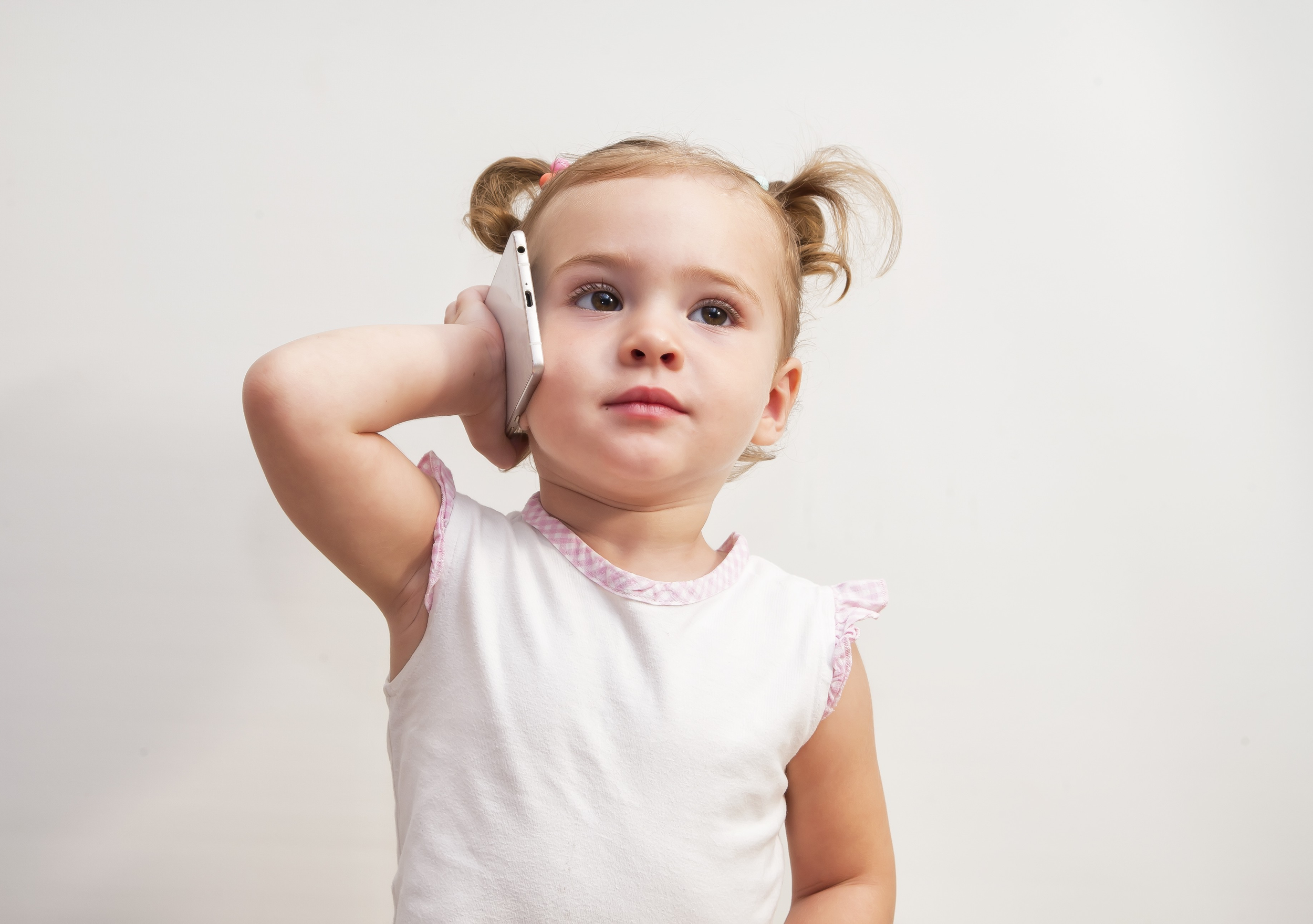 Young girl with curly hair in pigtails, holding a smartphone to her ear, wearing a white shirt with pink trim, standing against a plain white background.