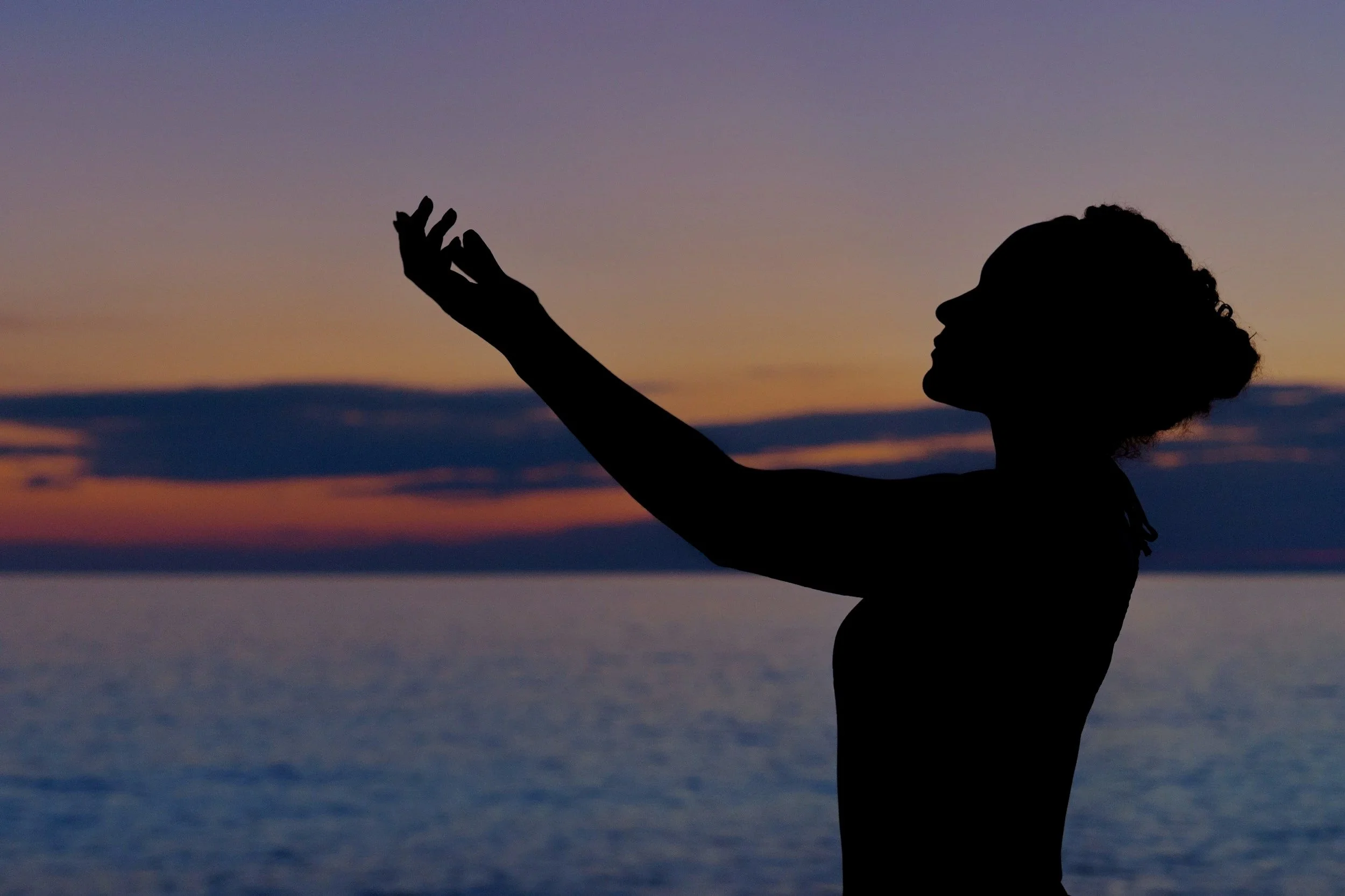 silhouette of a woman with curly hair reaching out her hand during sunset over the ocean
