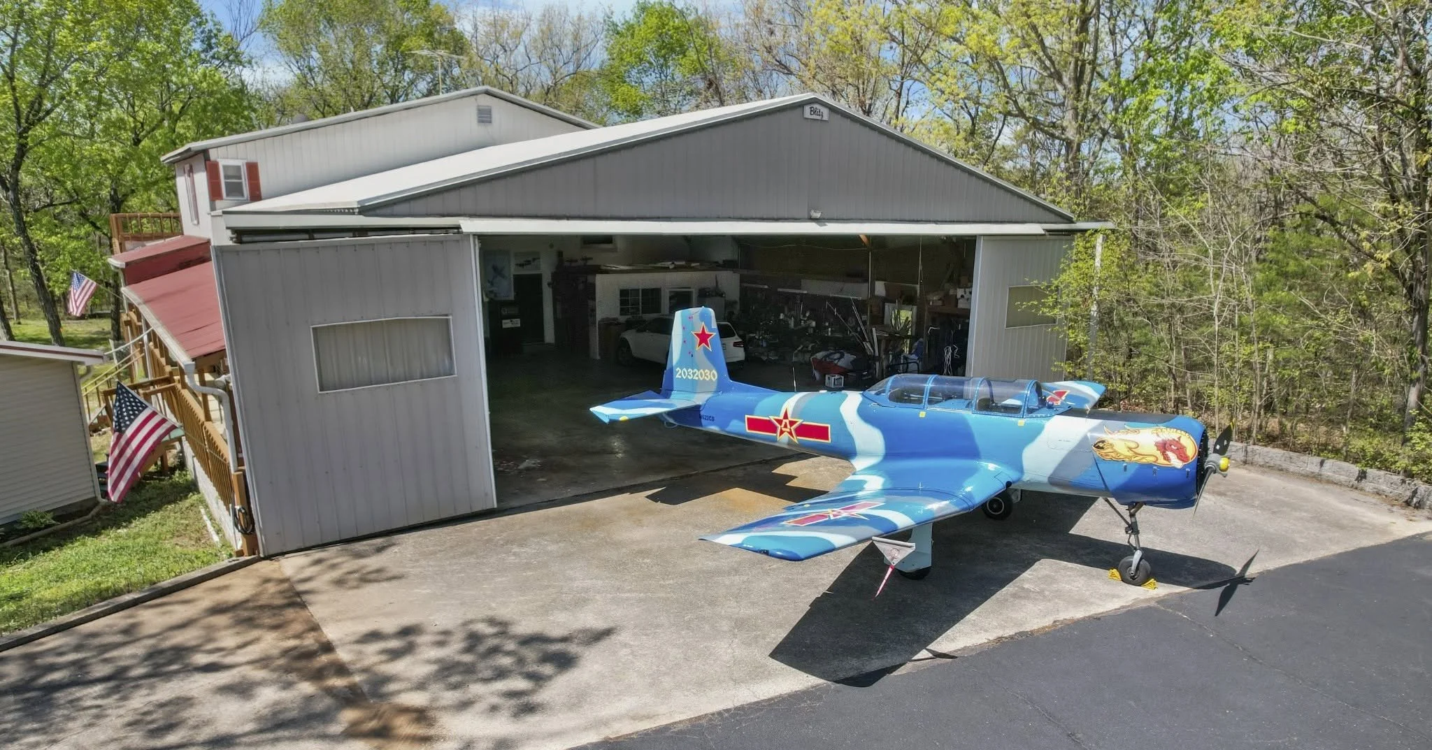 A small blue fighter jet with military insignia parked outside a garage, with trees and a fence with American flags nearby.