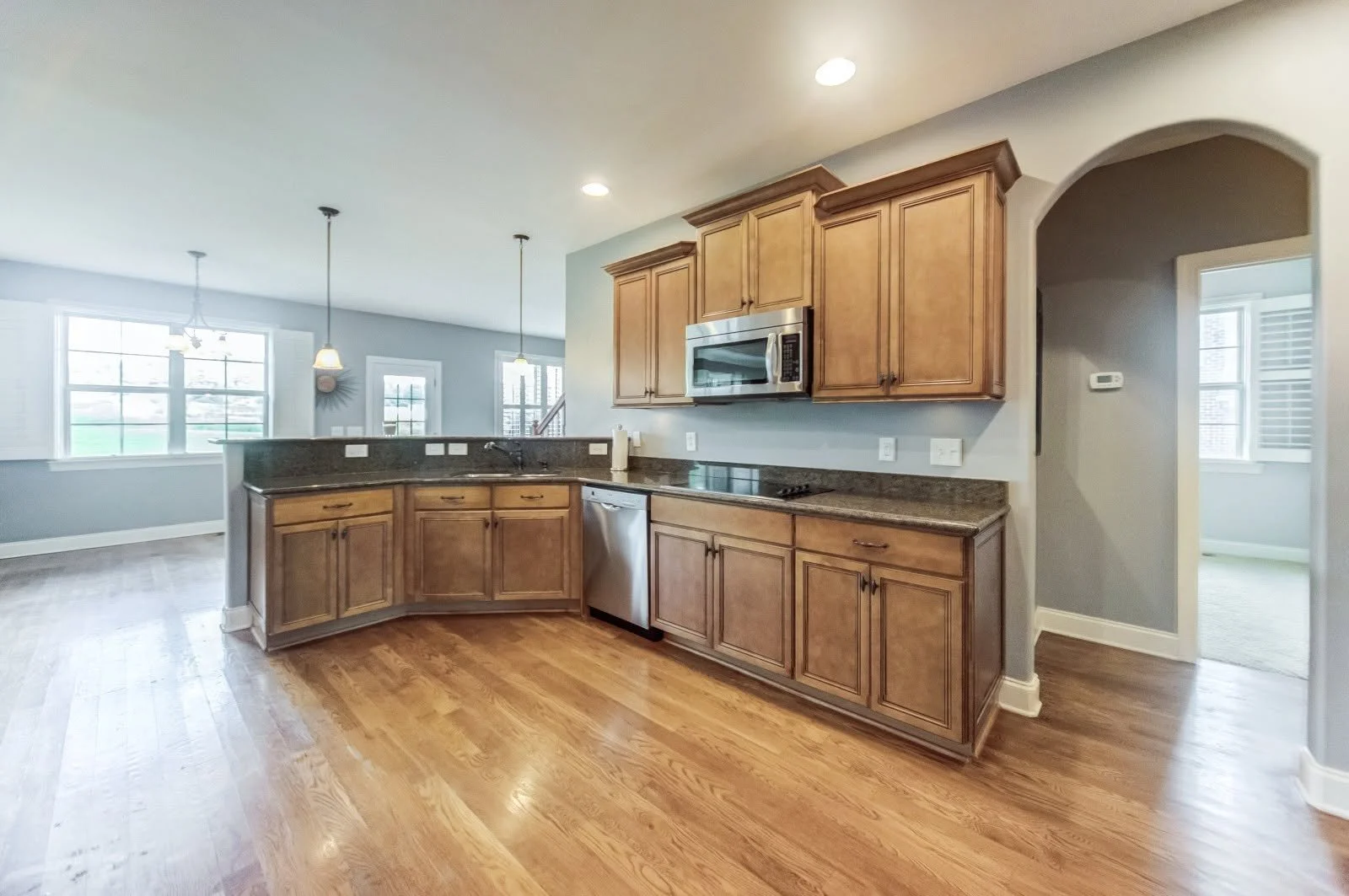 Kitchen with wooden cabinets, stainless steel microwave, granite countertops, hardwood floor, and an open living/dining area with large windows.