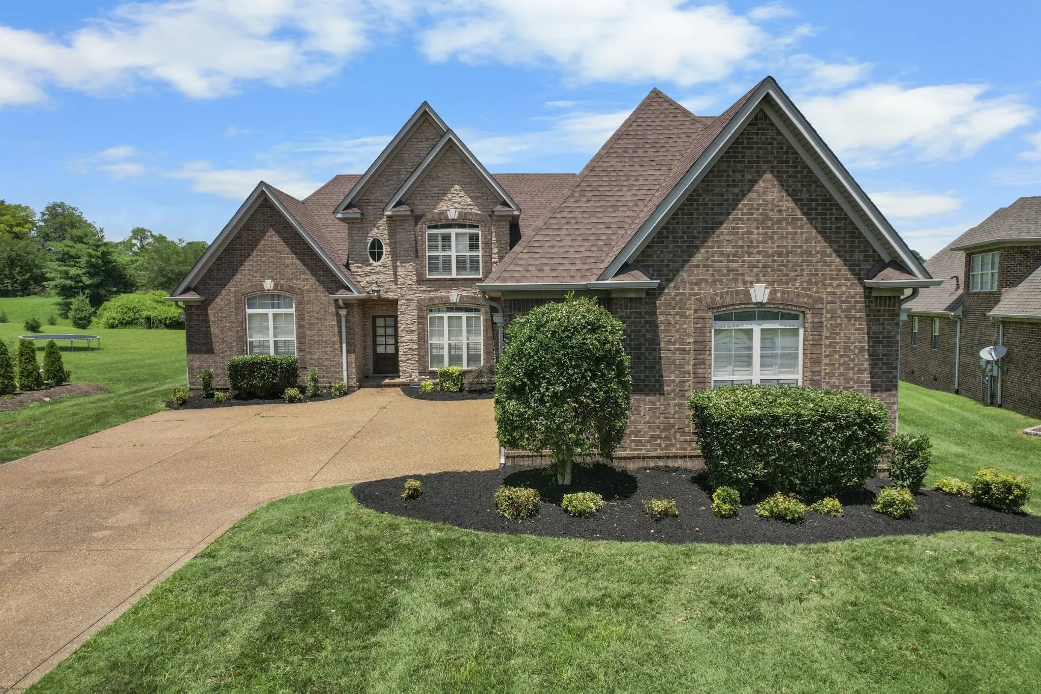 A two-story brick house with multiple gable roofs and large windows, surrounded by a well-manicured lawn, shrubs, and trees under a blue sky with some clouds.