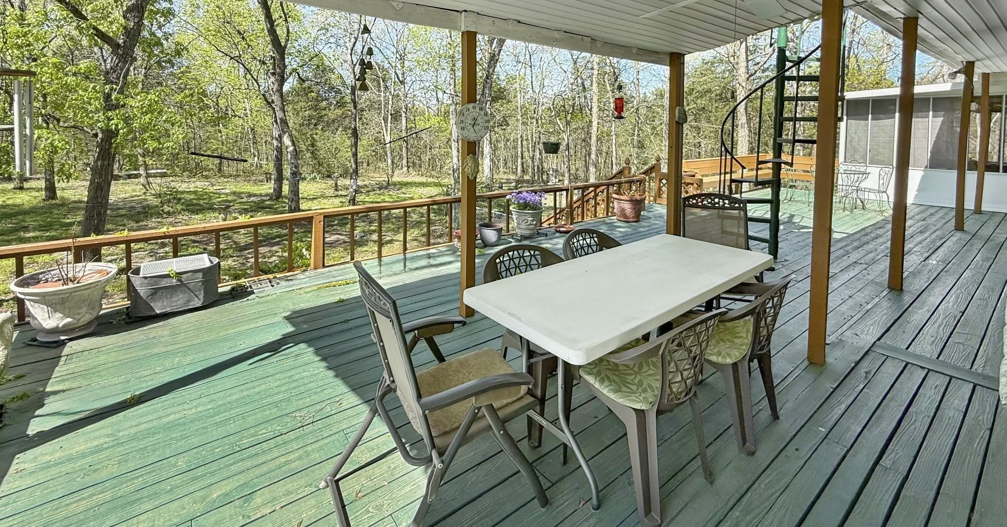 A spacious outdoor porch with wooden flooring, patio furniture, potted plants, a hanging clock, and a view of a wooded yard with trees and shaded area.