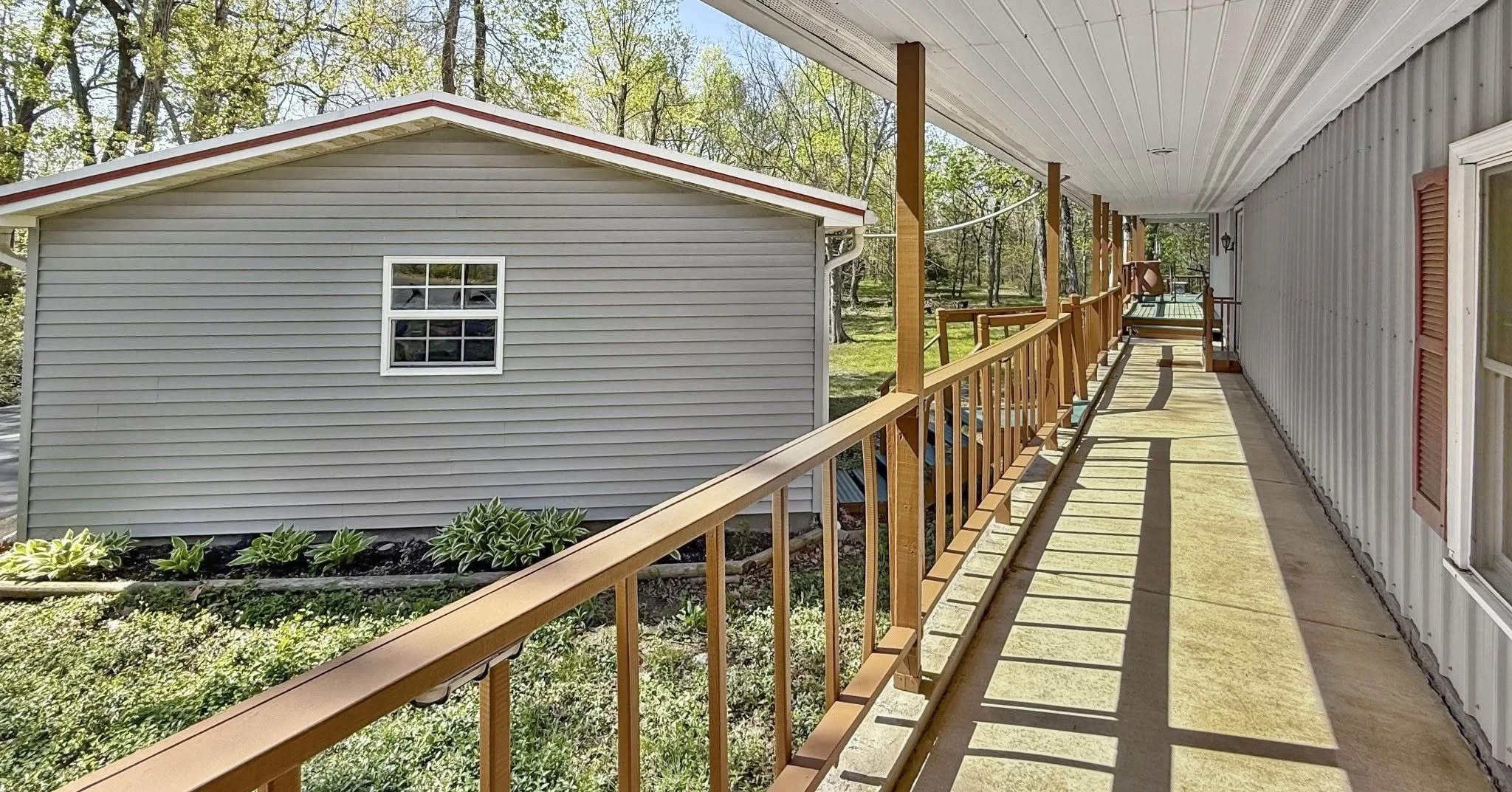 A view from a balcony or porch showing a long, narrow walkway with a yellow railing, beige flooring, and a white ceiling. The right side has the exterior wall of a house with red shutters and windows. The left side has a gray building with a small window, green plants, and trees in the background.
