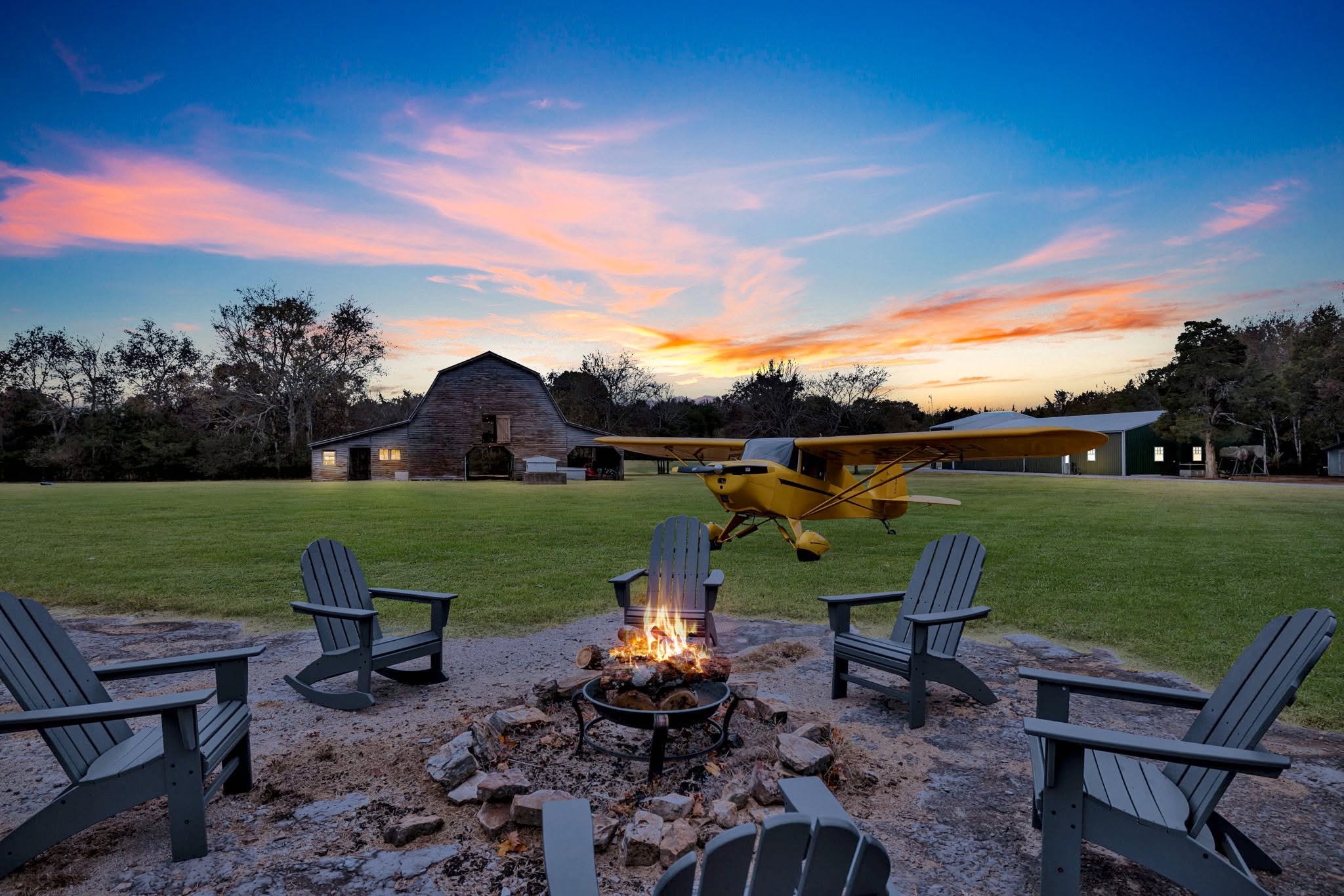 A backyard scene at sunset with a fire pit, surrounded by Adirondack chairs, a barn, and a small yellow airplane on the grass.