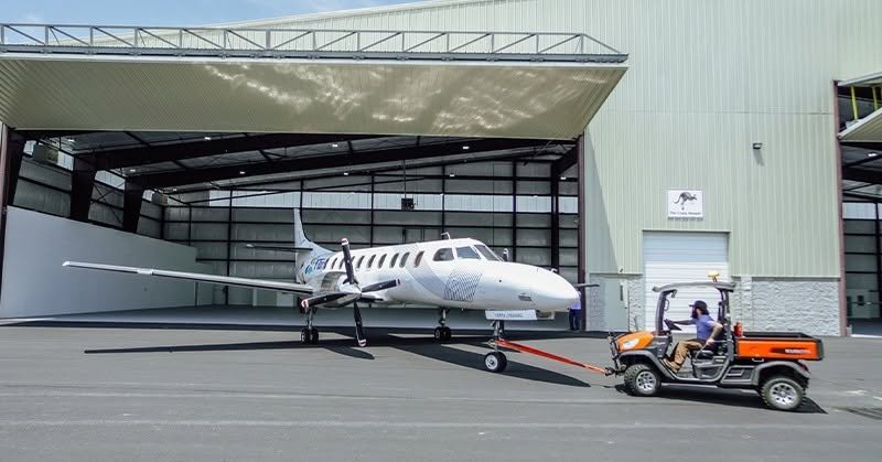 A small white private jet outside a hangar, tethered to a utility vehicle.