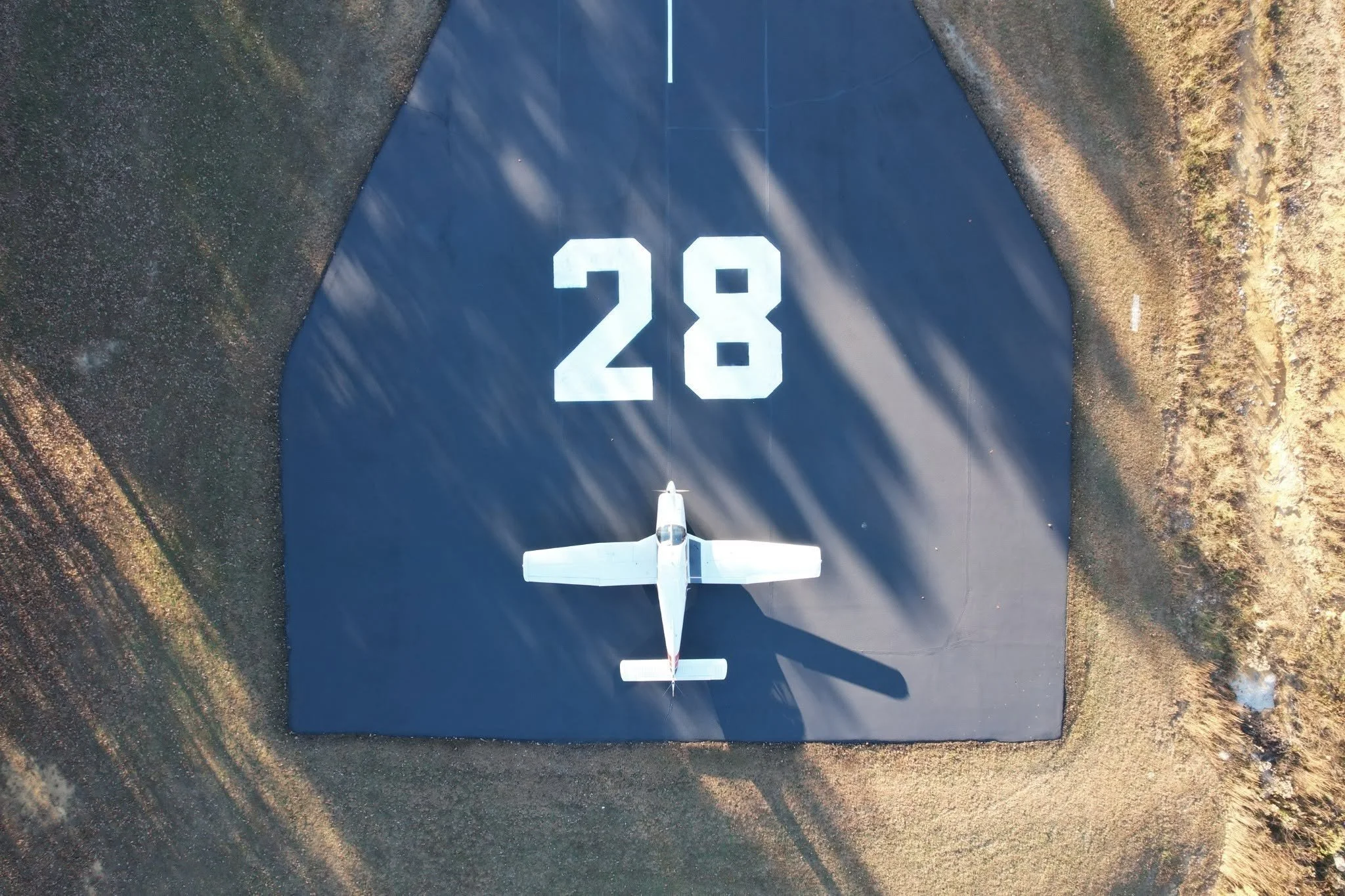 An airplane is positioned on a dark runway with white painted numbers '28'. The surrounding area is grassy with patches of dirt and gravel.