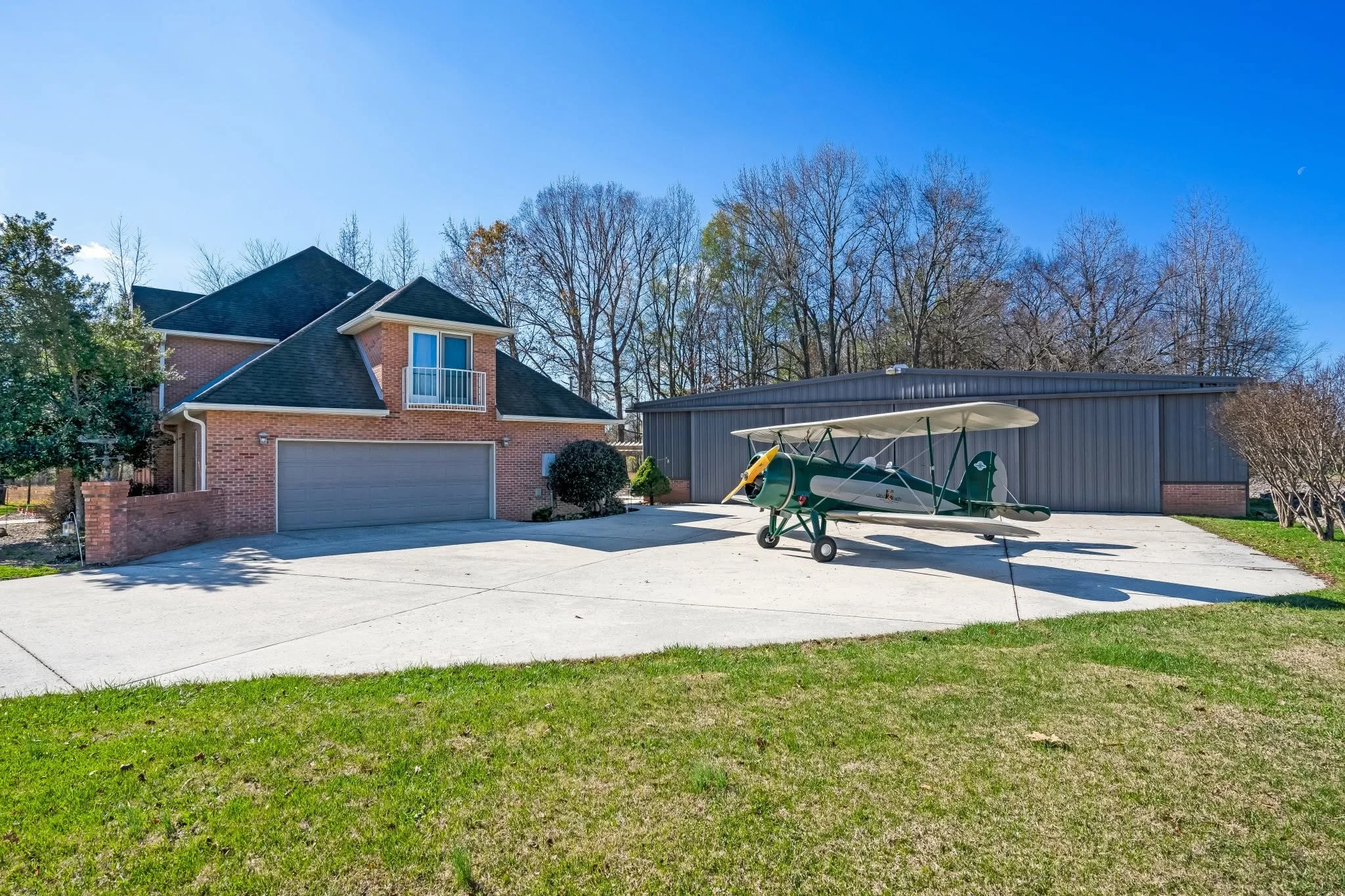 A suburban driveway with a small airplane parked in front of a brick house and a blue metal shed. The house has a two-car garage, a small balcony, and trees in the background. Clear blue sky overhead.