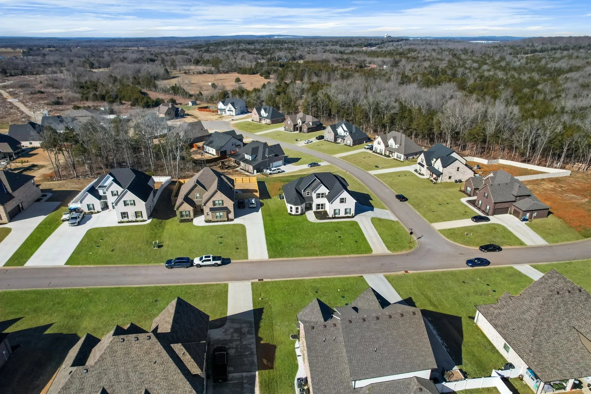 Aerial view of a suburban neighborhood with modern houses, green lawns, paved roads, and cars parked in driveways, surrounded by a wooded area and open fields.