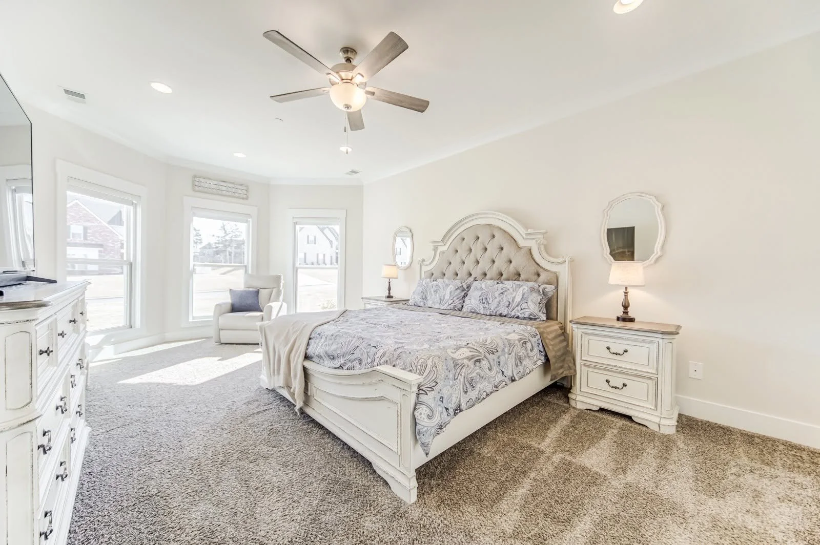 Bright bedroom with white furniture, a large bed with paisley bedding, bedside lamps, a white armchair, and multiple windows letting in natural light.