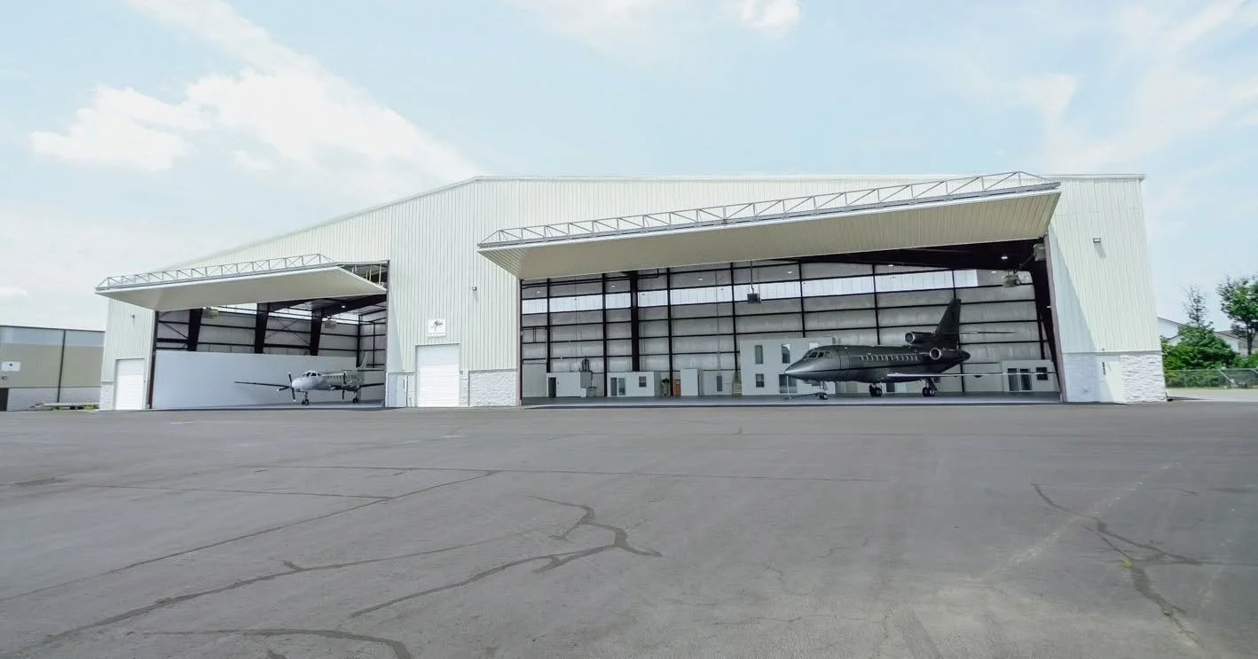 Large airplane hangar with two private jets inside, open on both sides, located on a paved area under a partly cloudy sky.