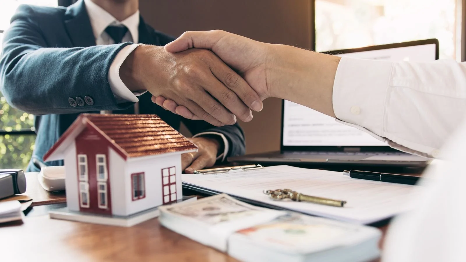 Two individuals in business attire shaking hands across a desk with a small model house, money, and legal documents.
