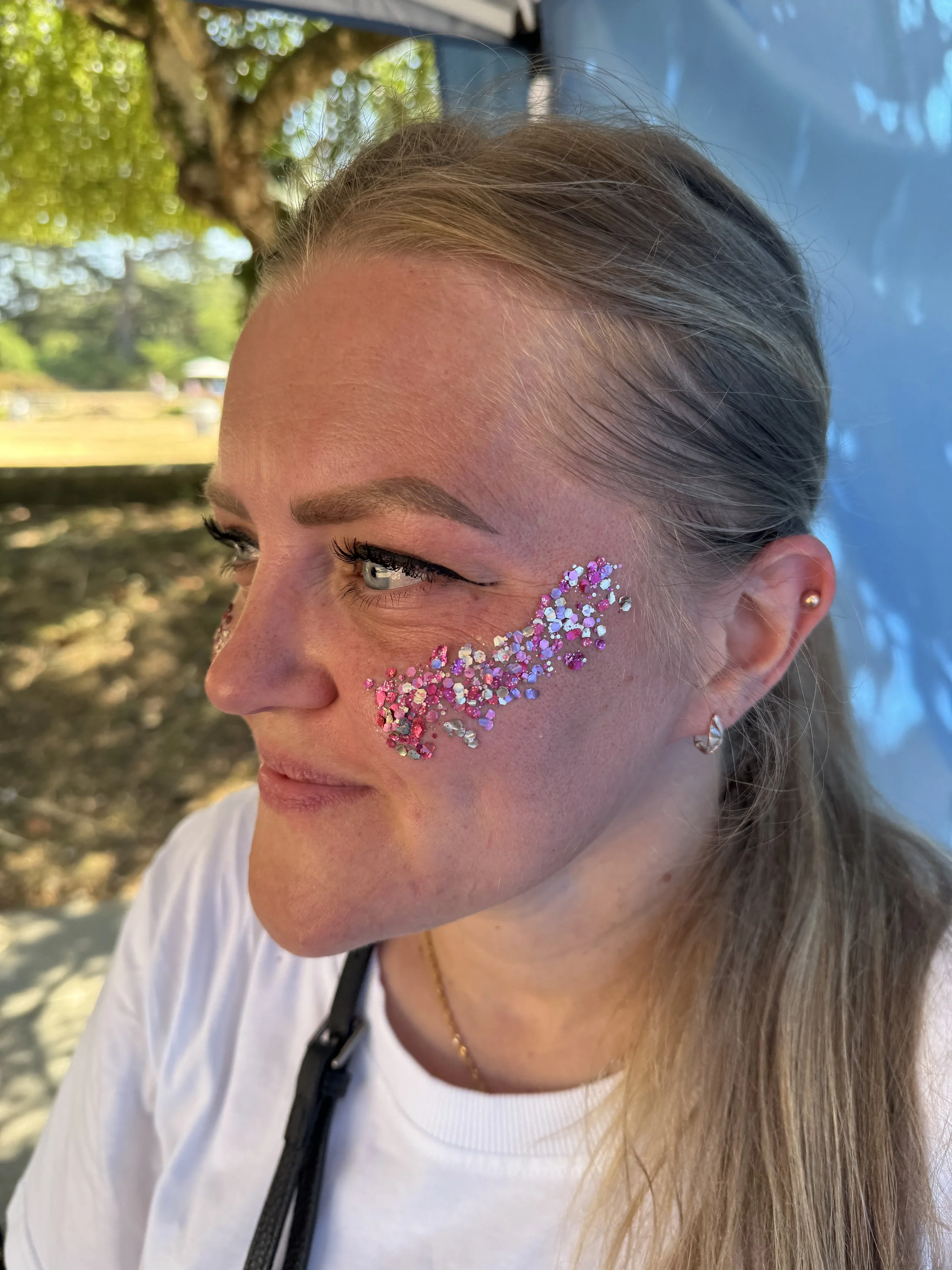 Close-up of a woman's face with glittery pink and white rhinestones applied around her eye, outdoors with trees and a blue tent in the background.