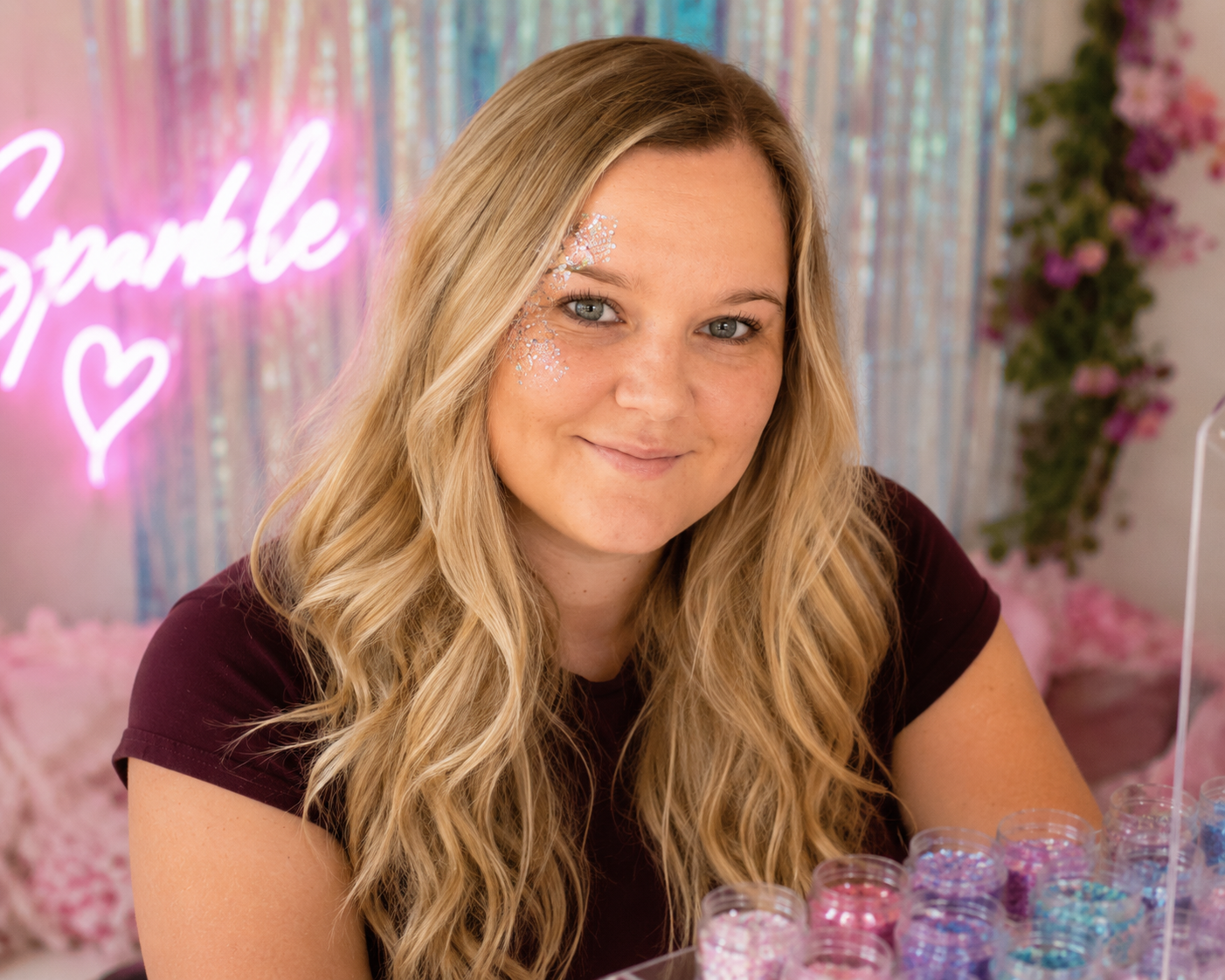 A woman with long, wavy blonde hair and light makeup, smiling at the camera, with glitter on her face, in front of a colorful background with a pink neon sign that says 'Sparkle' and pink flowers.