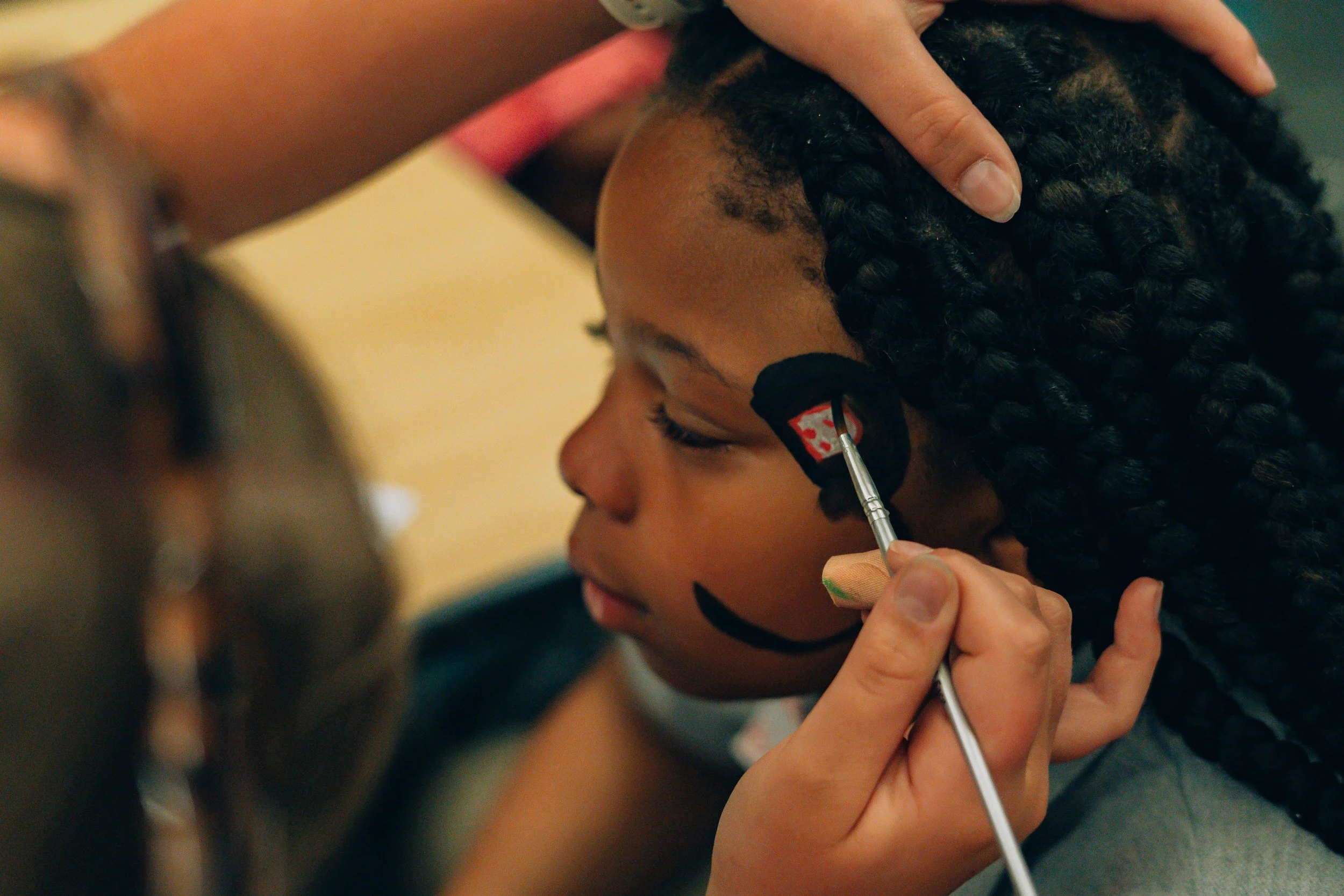 A young girl with curly hair has her face painted and is getting her face painted by an artist. The artist is holding a thin paintbrush near the girl’s cheek.