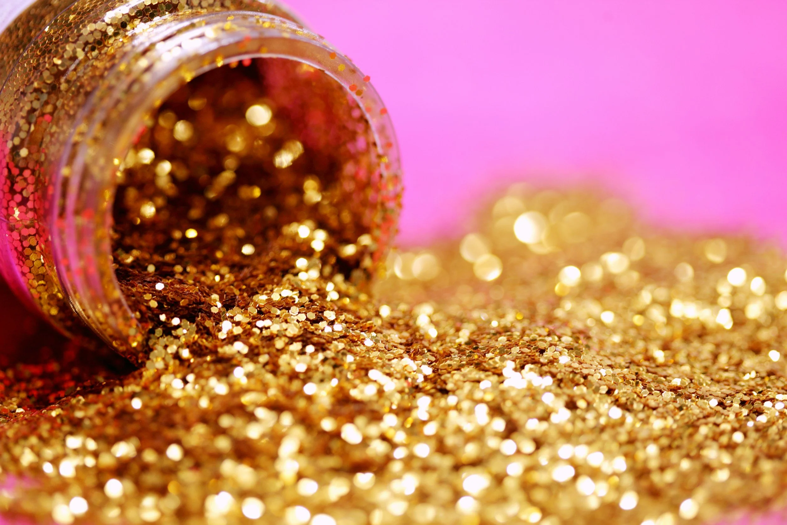Gold glitter spilled from a jar onto a pink surface.