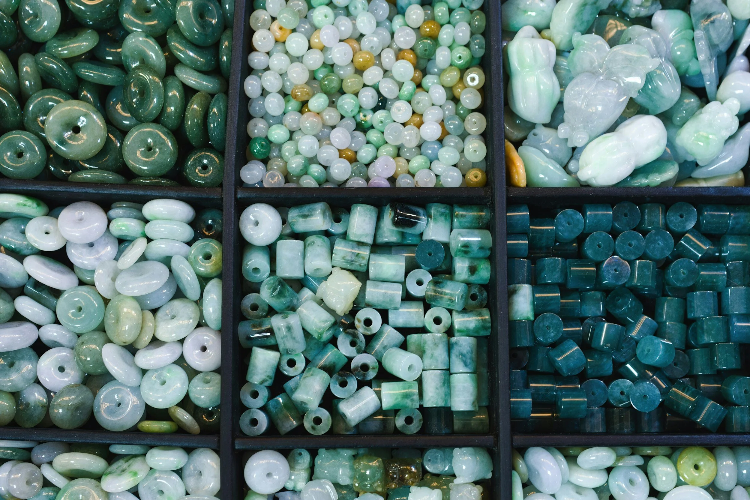Display of various green and white beads made of jade or similar material, organized in black trays.