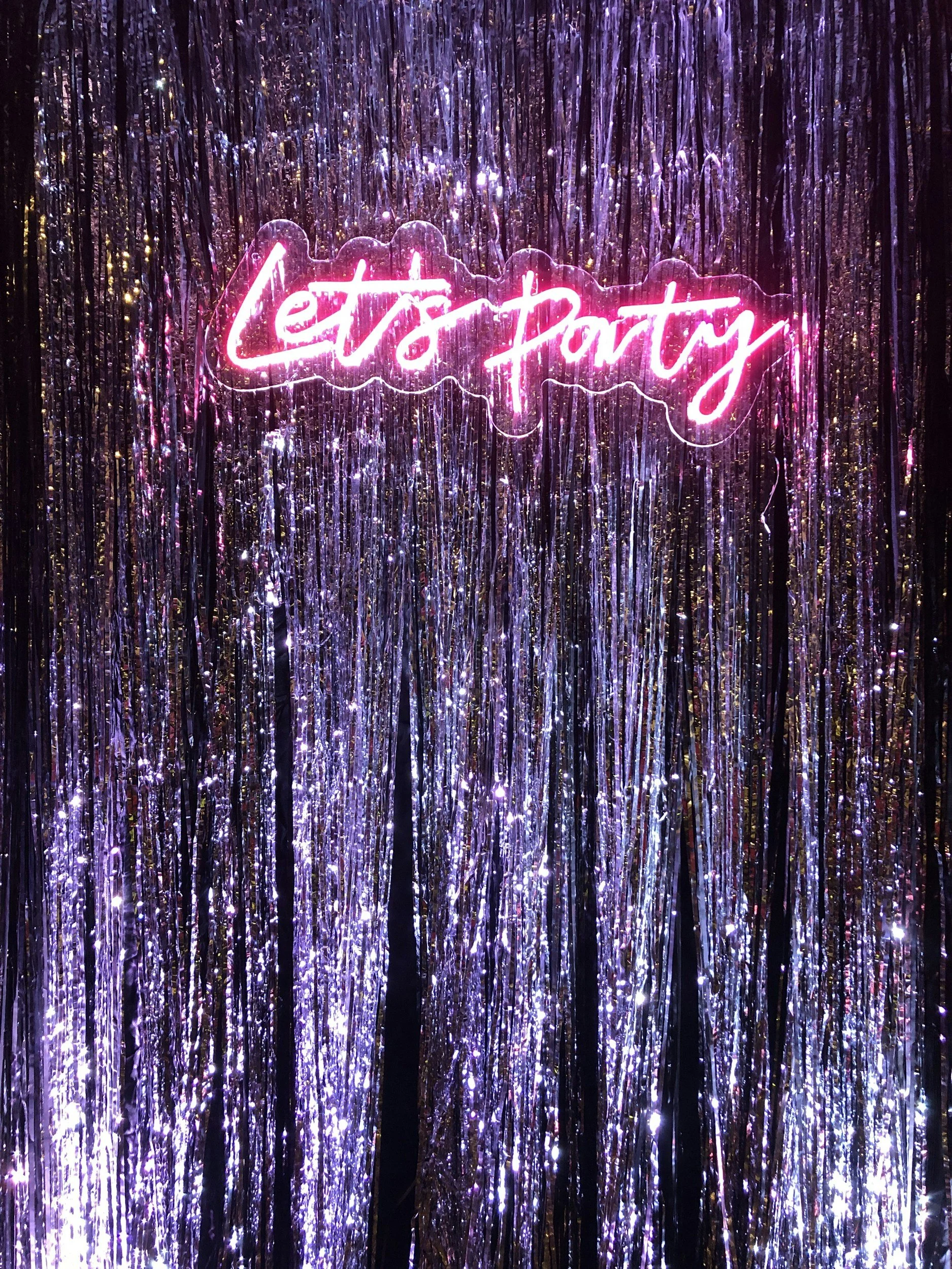 Neon sign reading 'Let’s Party' in pink, mounted on shimmering purple and holographic tinsel background.