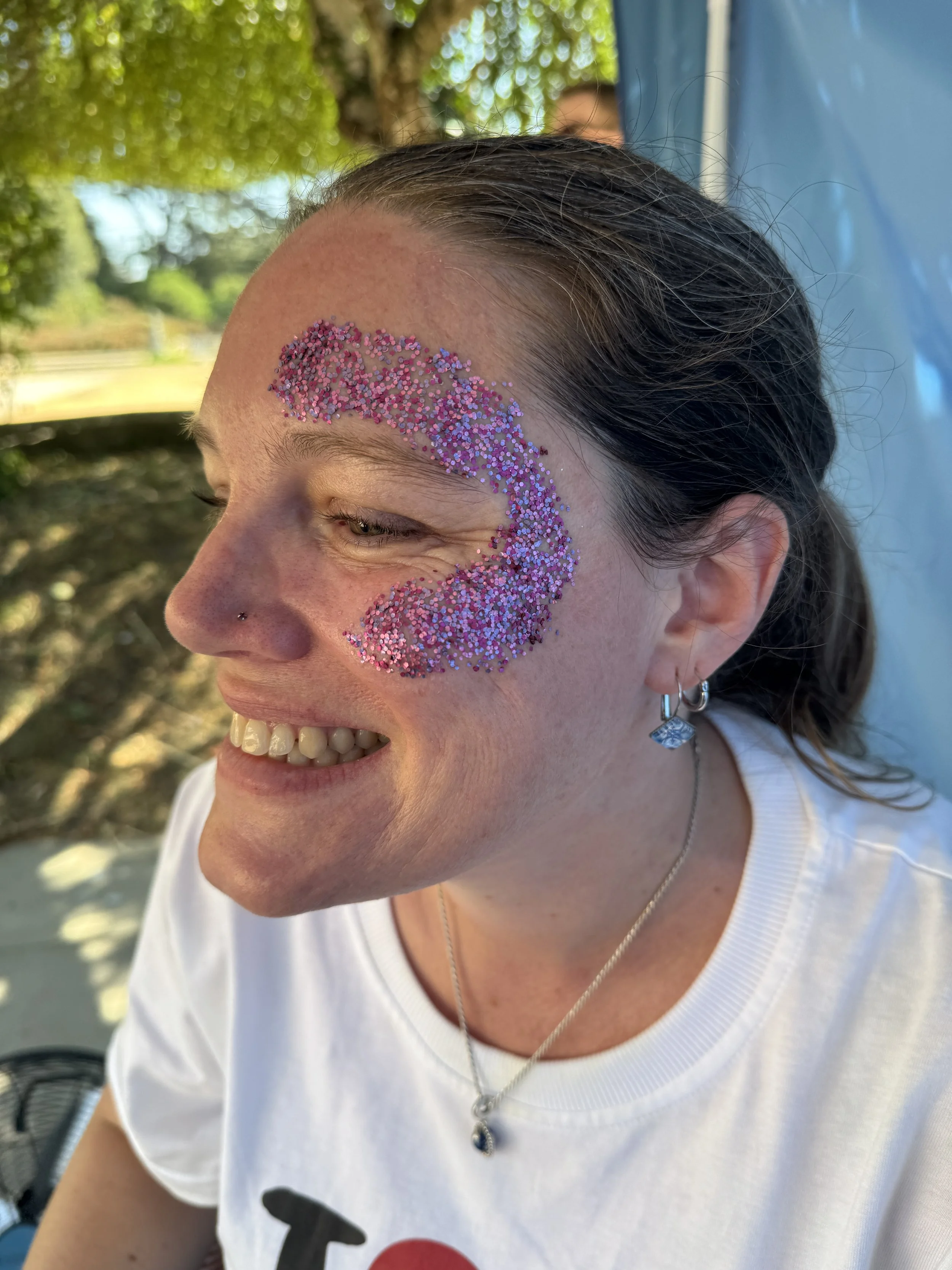 A woman with dark hair and earrings smiling, with pink and purple glitter on her forehead, outdoors in sunlight with blurred trees in the background.