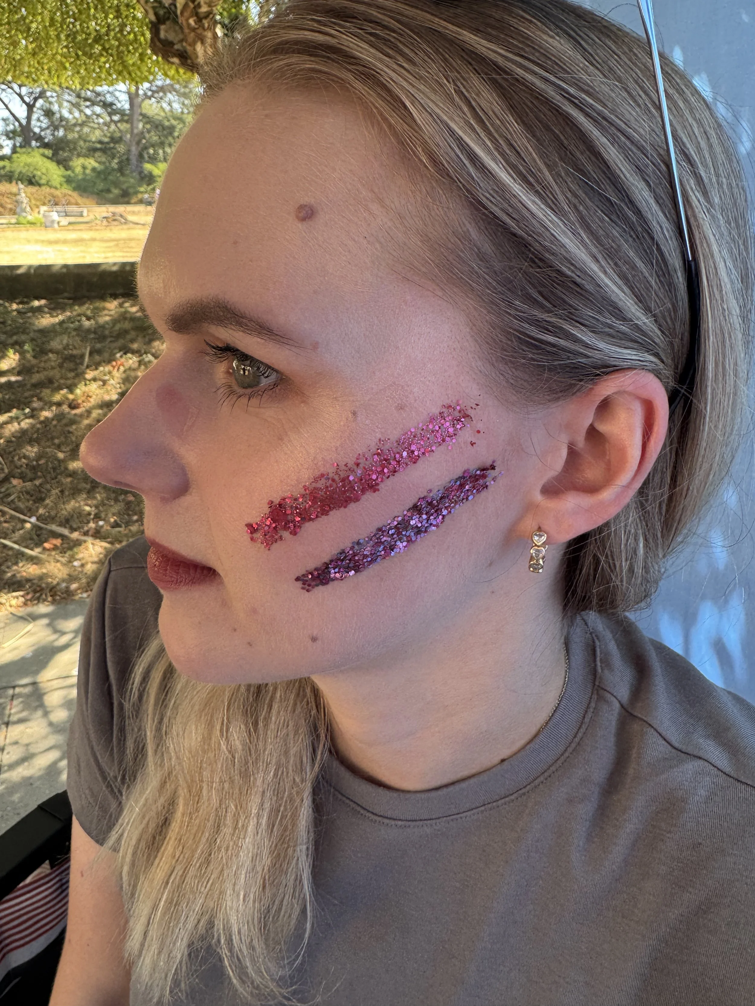 A close-up of a woman with light brown hair, wearing glitter makeup stripes on her cheek and a small earring. The photo is taken outdoors, with trees and sky in the background.