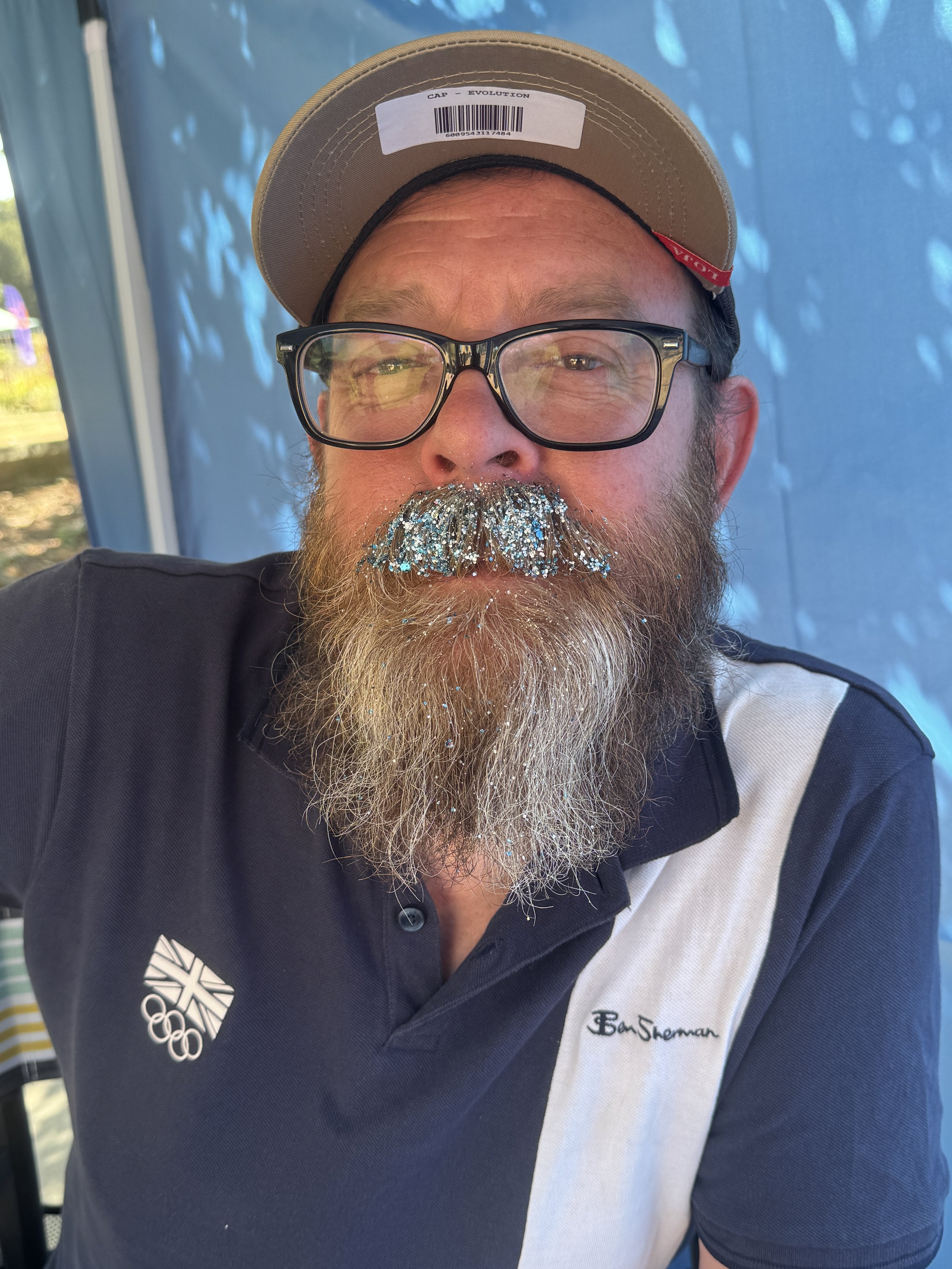 Close-up of a man wearing glasses, a tan cap with a barcode label, and a shirt with the Ben Sherman logo and Olympic emblem, with a blue background.