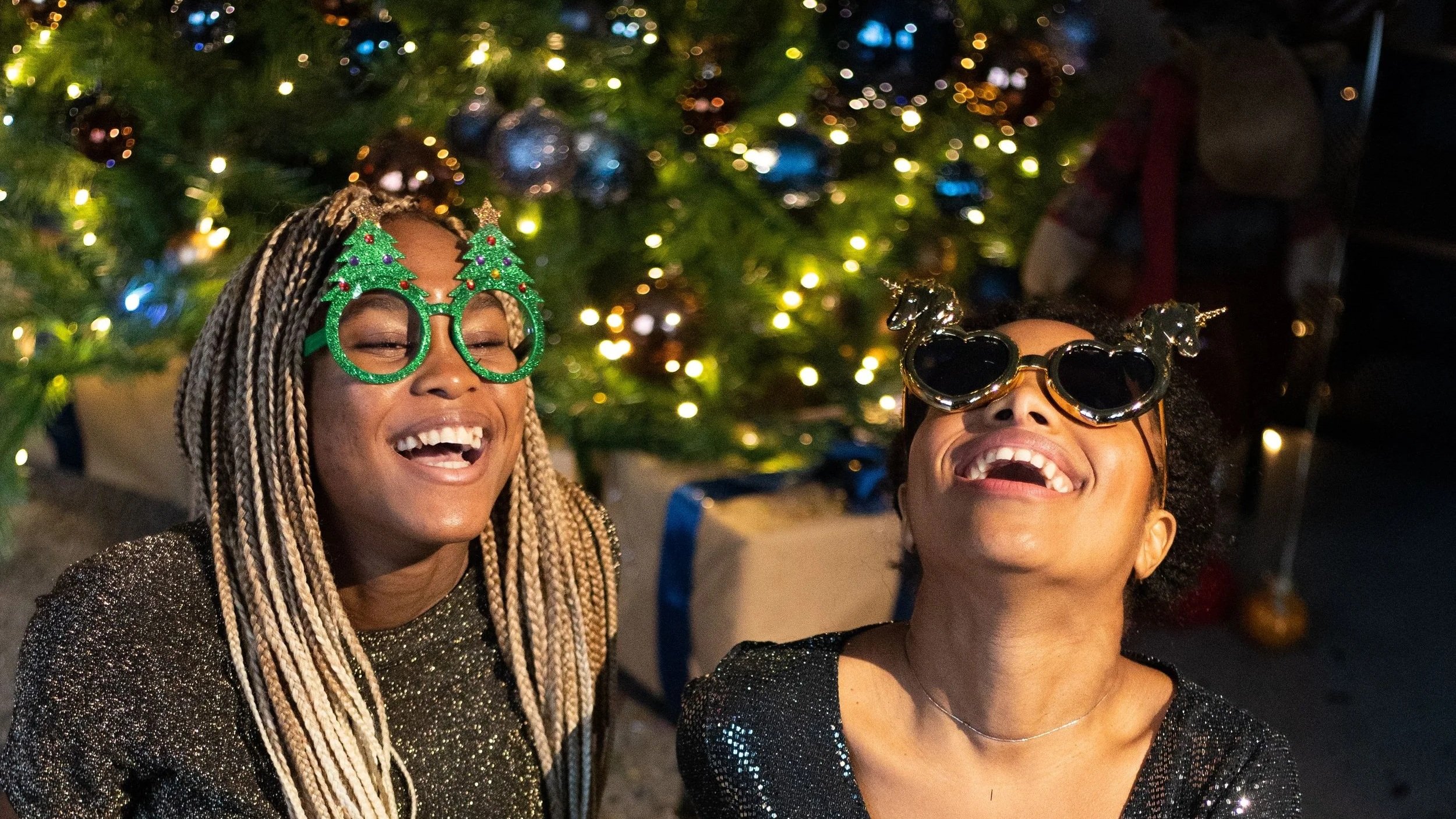 Two women wearing festive holiday glasses laughing in front of a decorated Christmas tree.