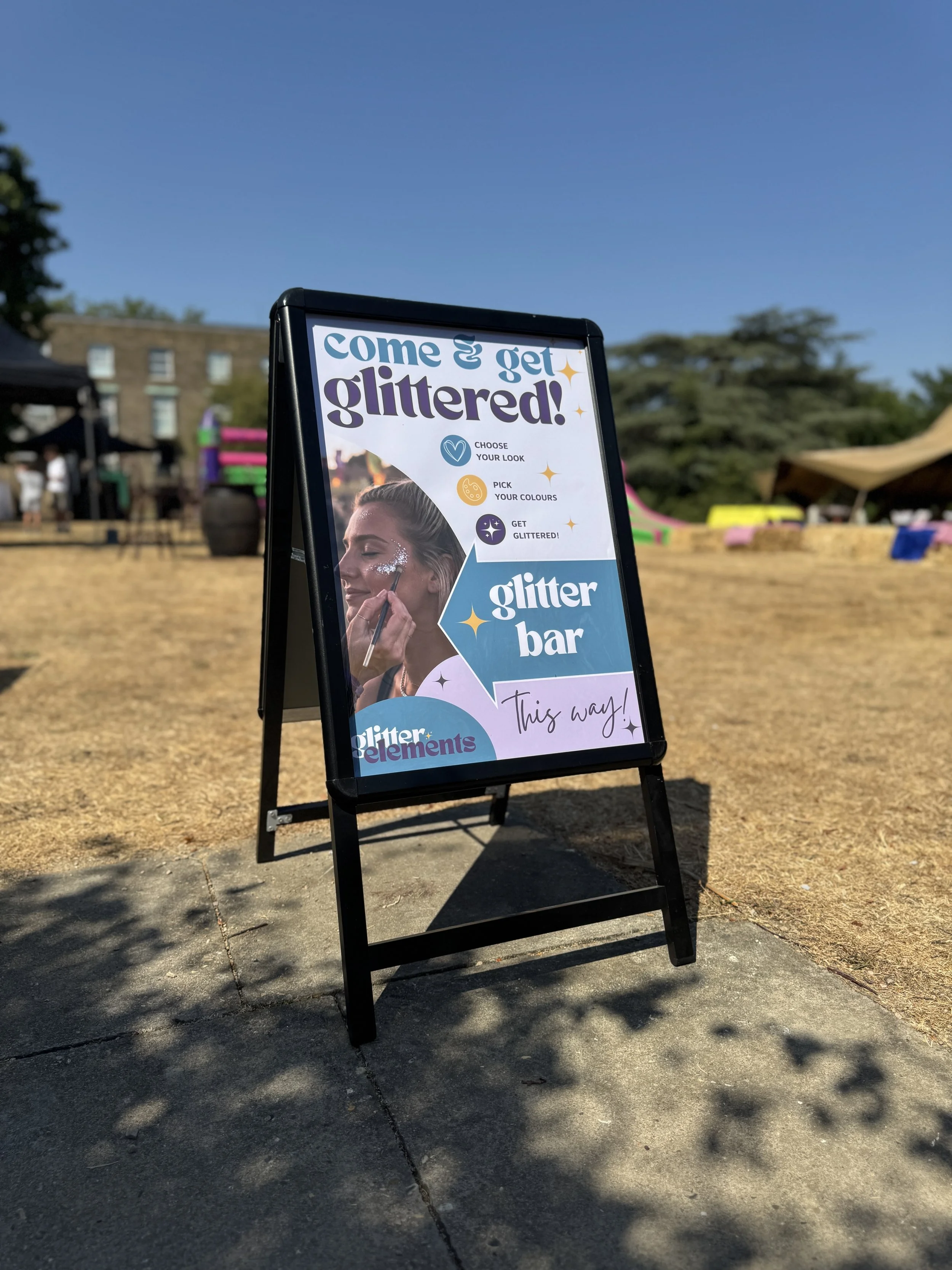 Outdoor sign advertising a glitter bar event offering options to choose look and colors for glitter elements, with a colorful background and people at a fair setting.
