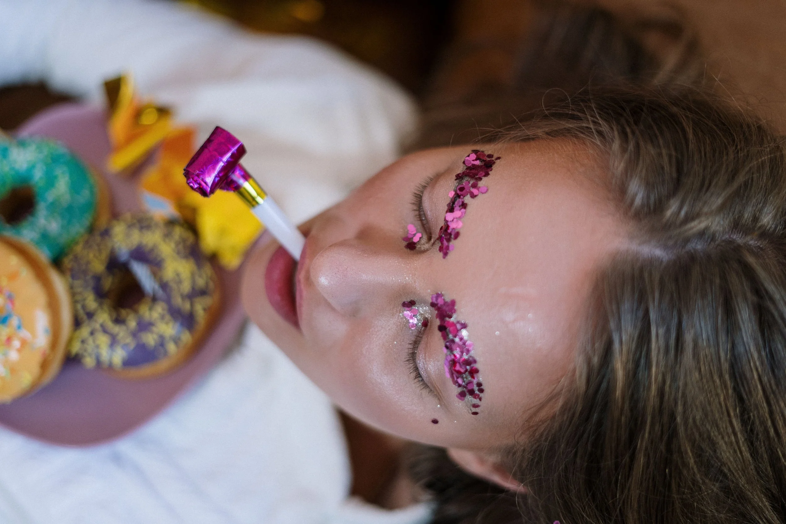 A woman is lying down with her eyes closed, decorated with pink glitter on her face and a small pink and purple party horn in her mouth. There are colorful donuts with frosting and sprinkles visible in the foreground.