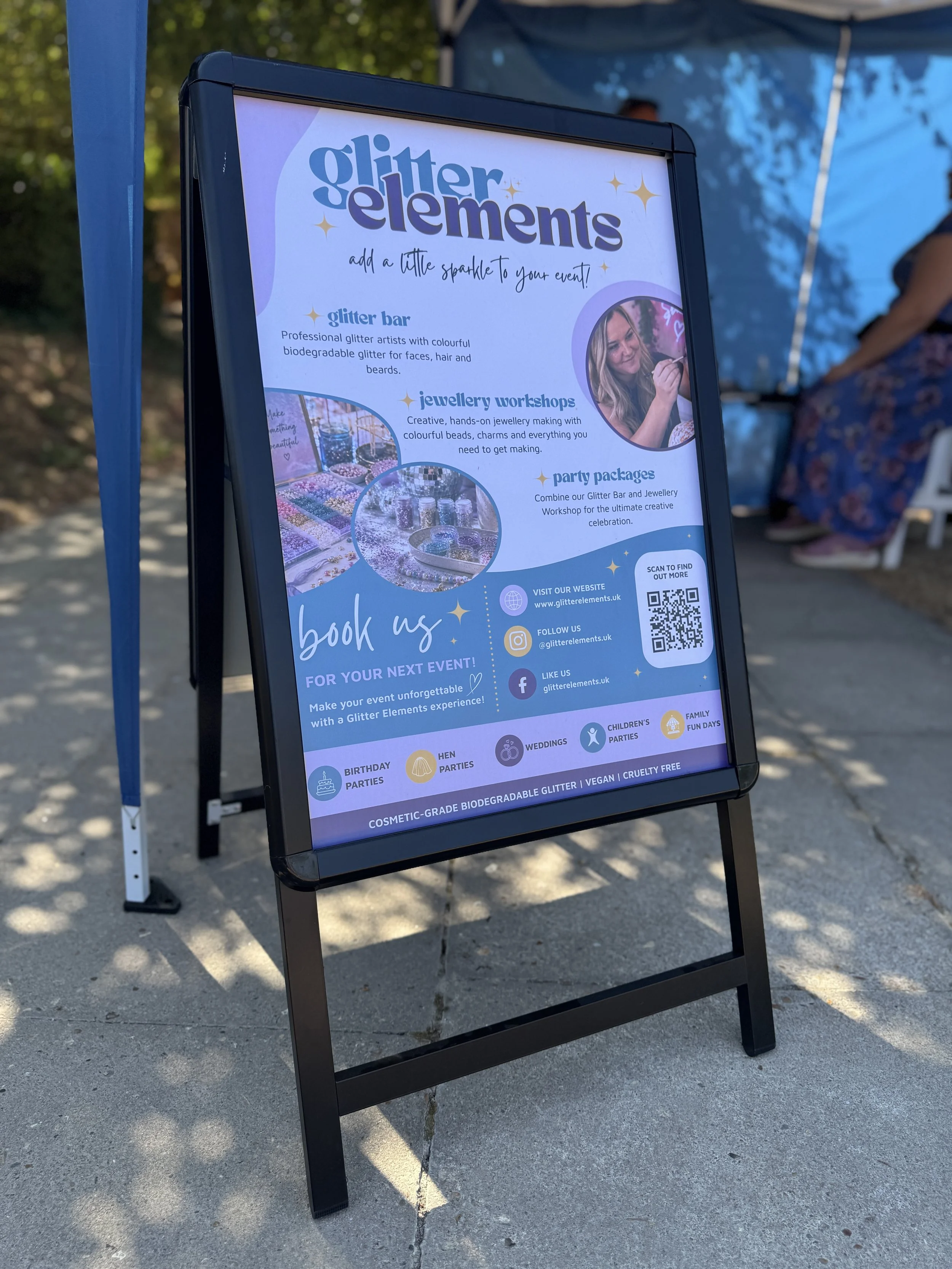 A black stand-up signboard with a colorful flyer for Glitter Elements, an event business offering glitter bar, jewelry workshops, and party packages. The flyer includes images of glitter and jewelry making supplies, with social media icons and a QR c